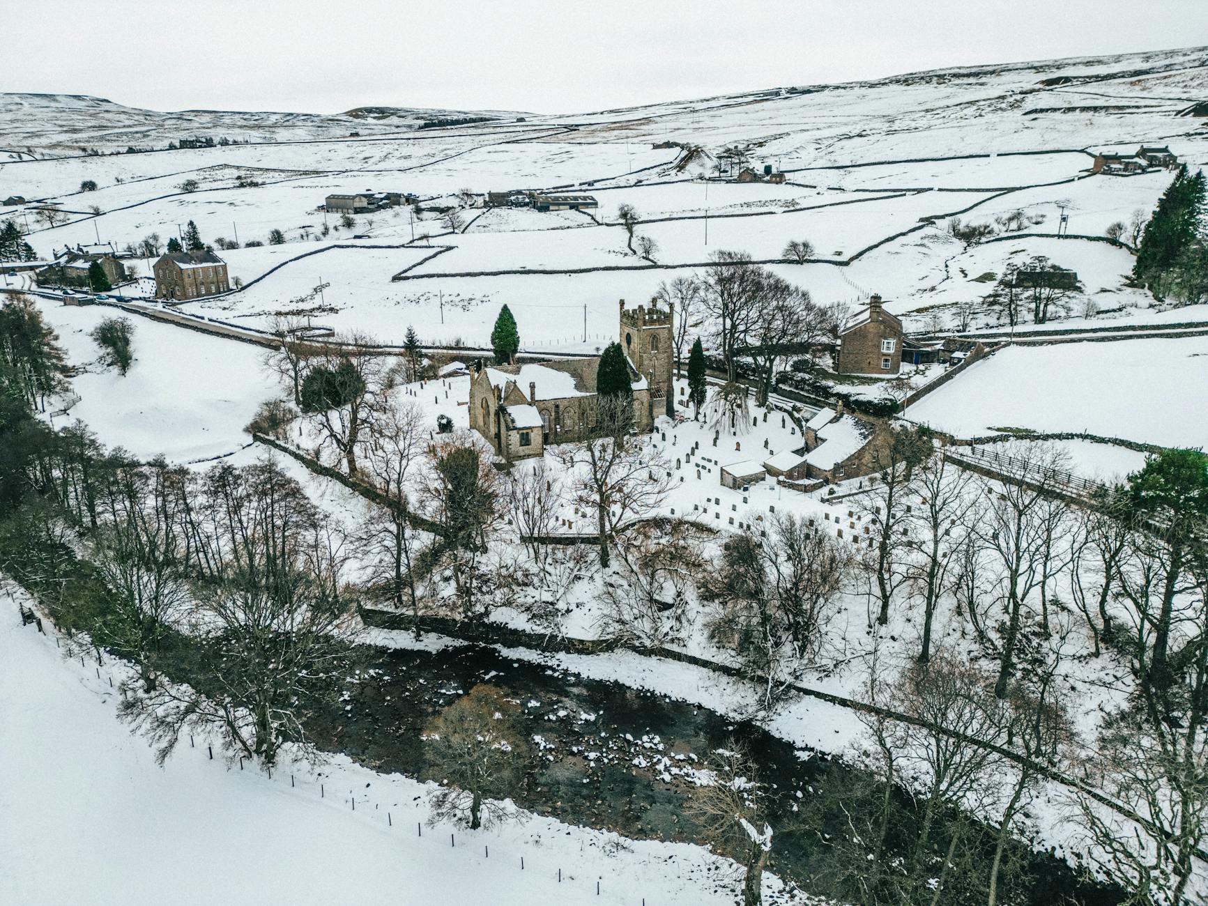 An aerial view of a snow-covered Cotswold village with a church and river in winter