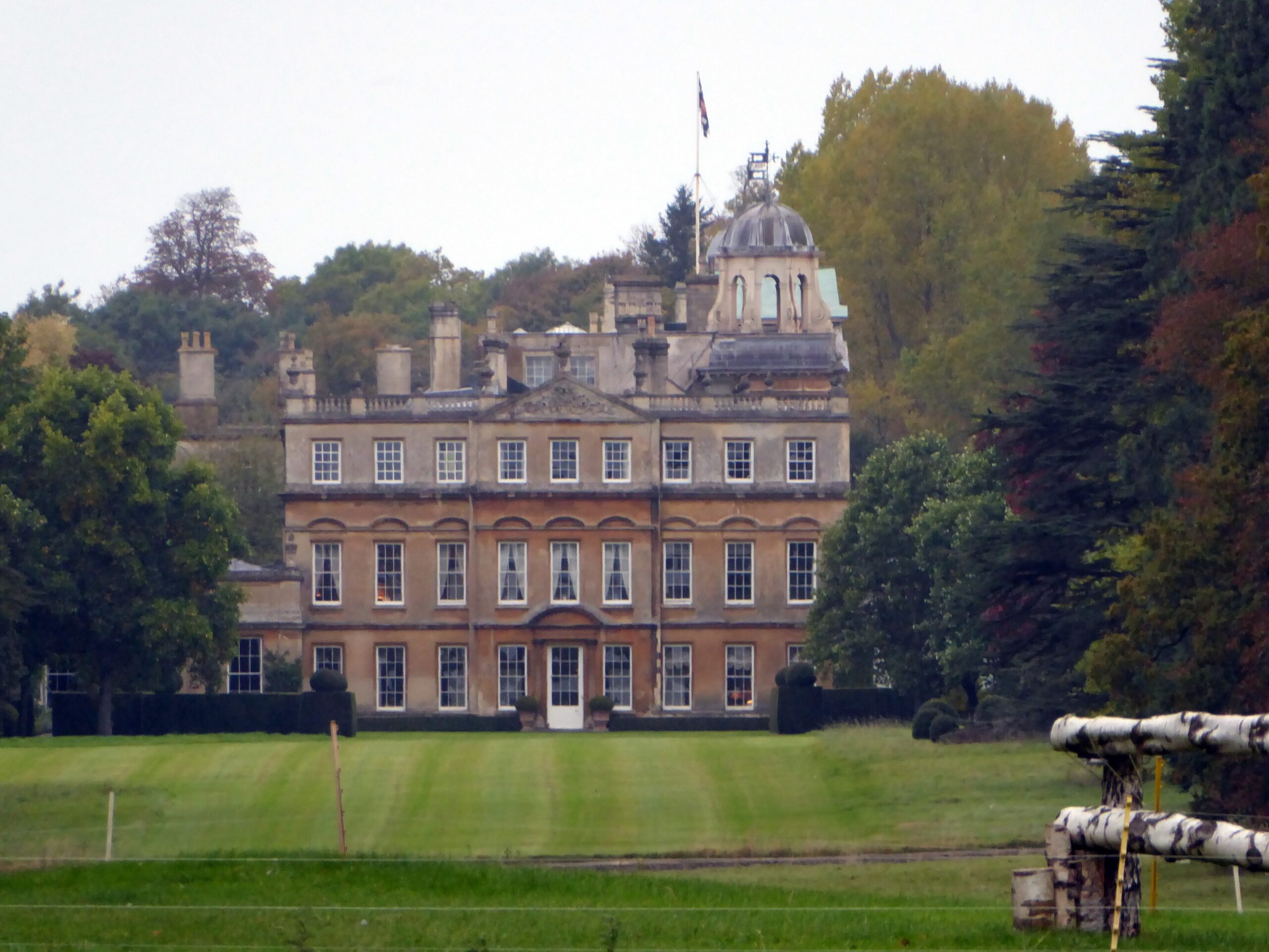 The Palladian north front of Badminton House in Gloucestershire, with a domed cupola and flag above, seen across mown parkland with a timber cross-country fence in the foreground