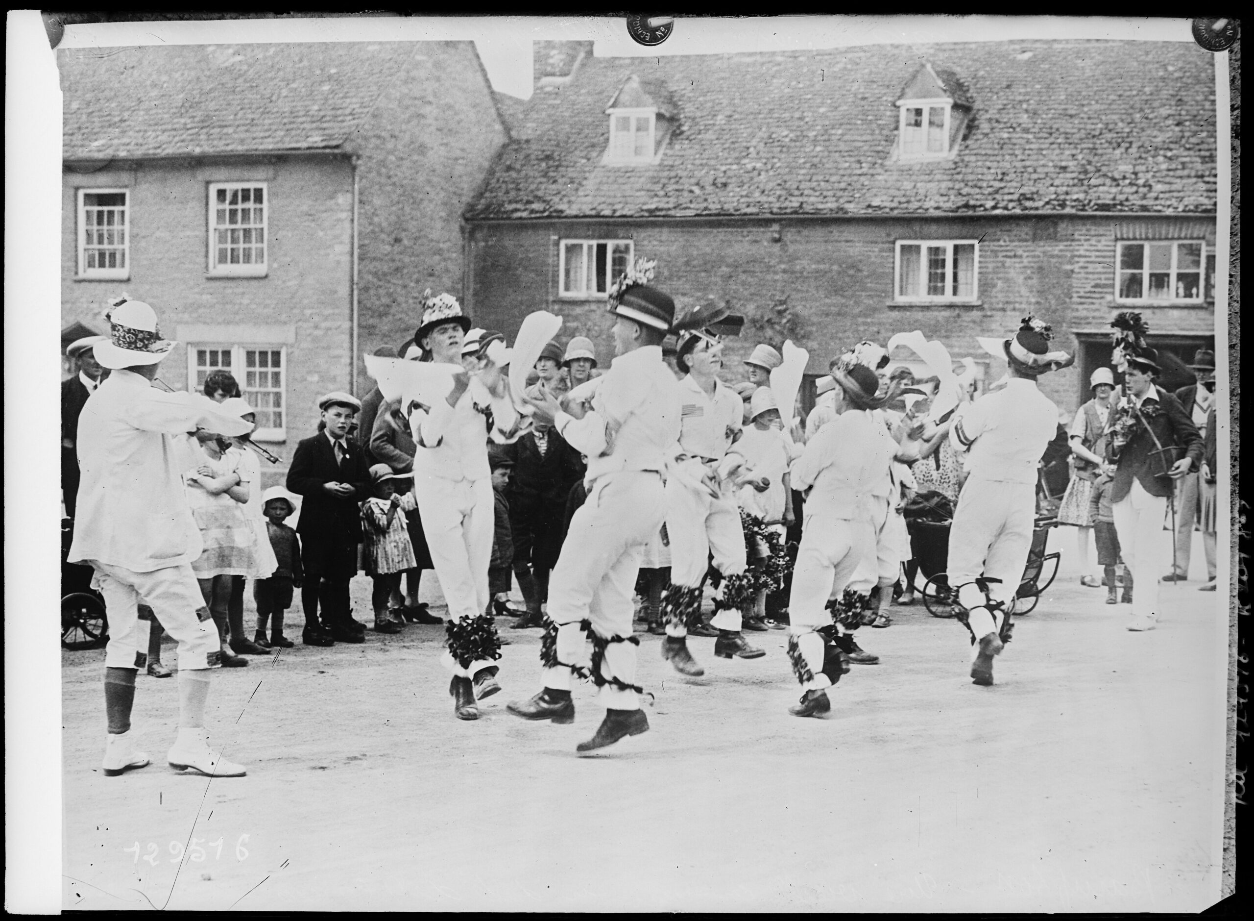A black-and-white press photograph from the 1920s showing Bampton Morris dancers in white with flowered hats and bells at their knees, dancing in the street watched by a crowd of villagers and children
