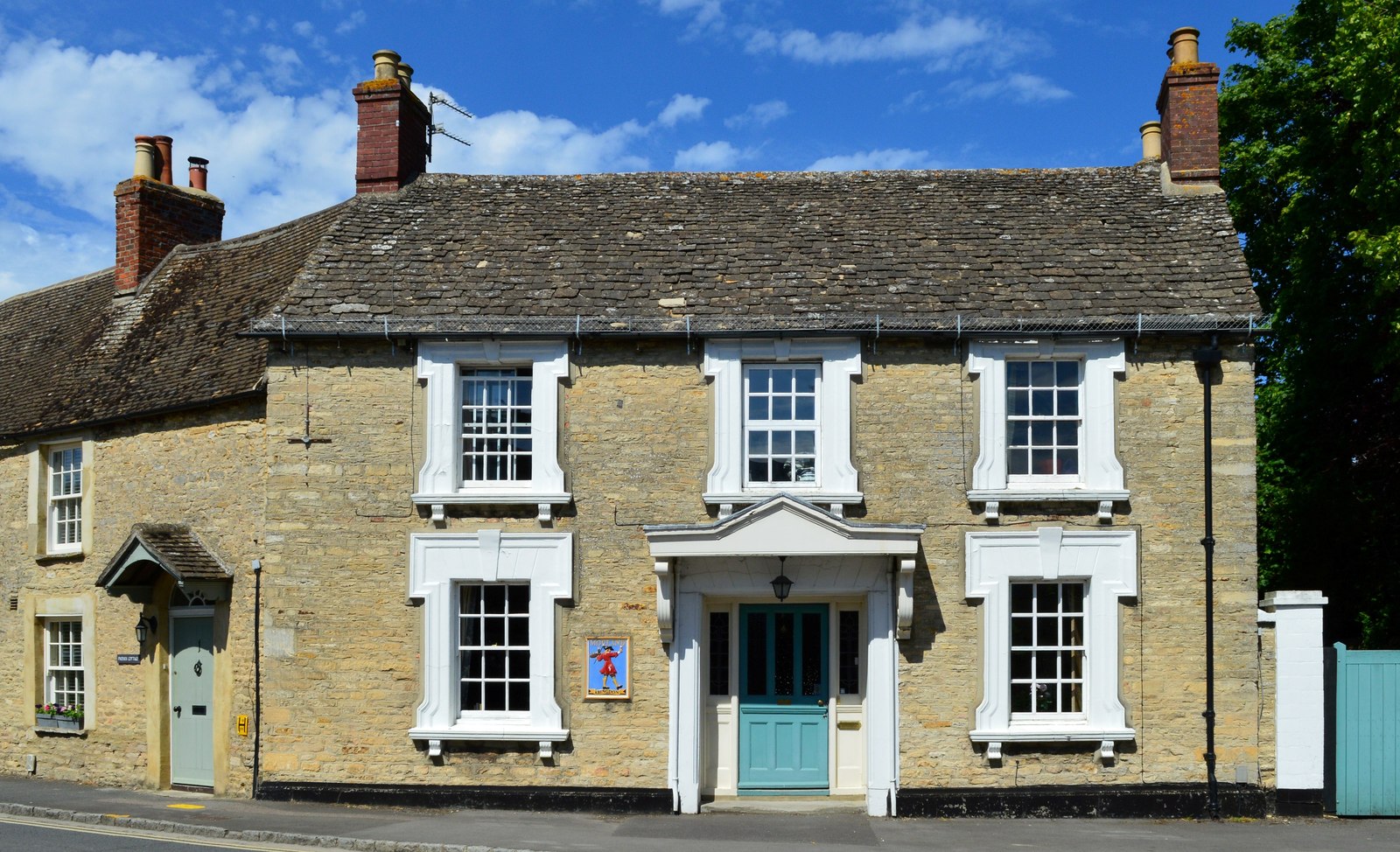 The Old George in Bampton, a two-storey stone house with a stone-tiled roof, white window surrounds, a pedimented porch with a teal door and a painted sign of a figure in a red coat