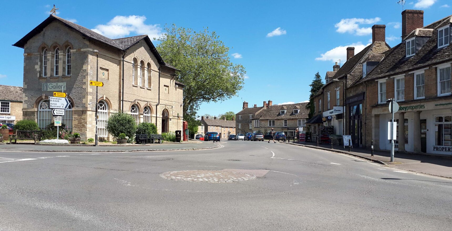Bampton's market square on a sunny day, with the arched-windowed stone town hall housing a gallery on the left and a row of stone shops and houses on the right