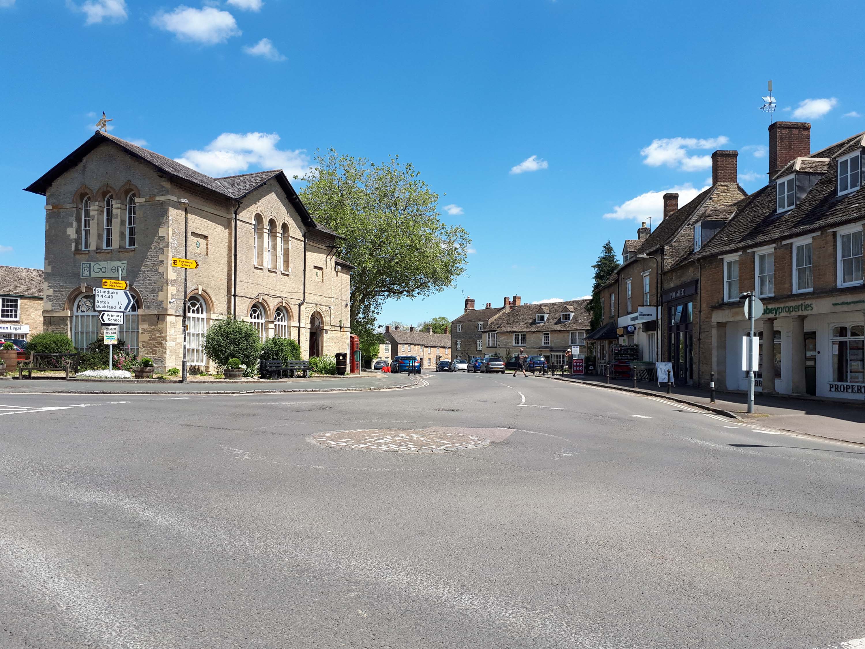 Bampton's market square on a sunny day, with the arched-windowed stone town hall housing a gallery on the left and a row of stone shops and houses on the right