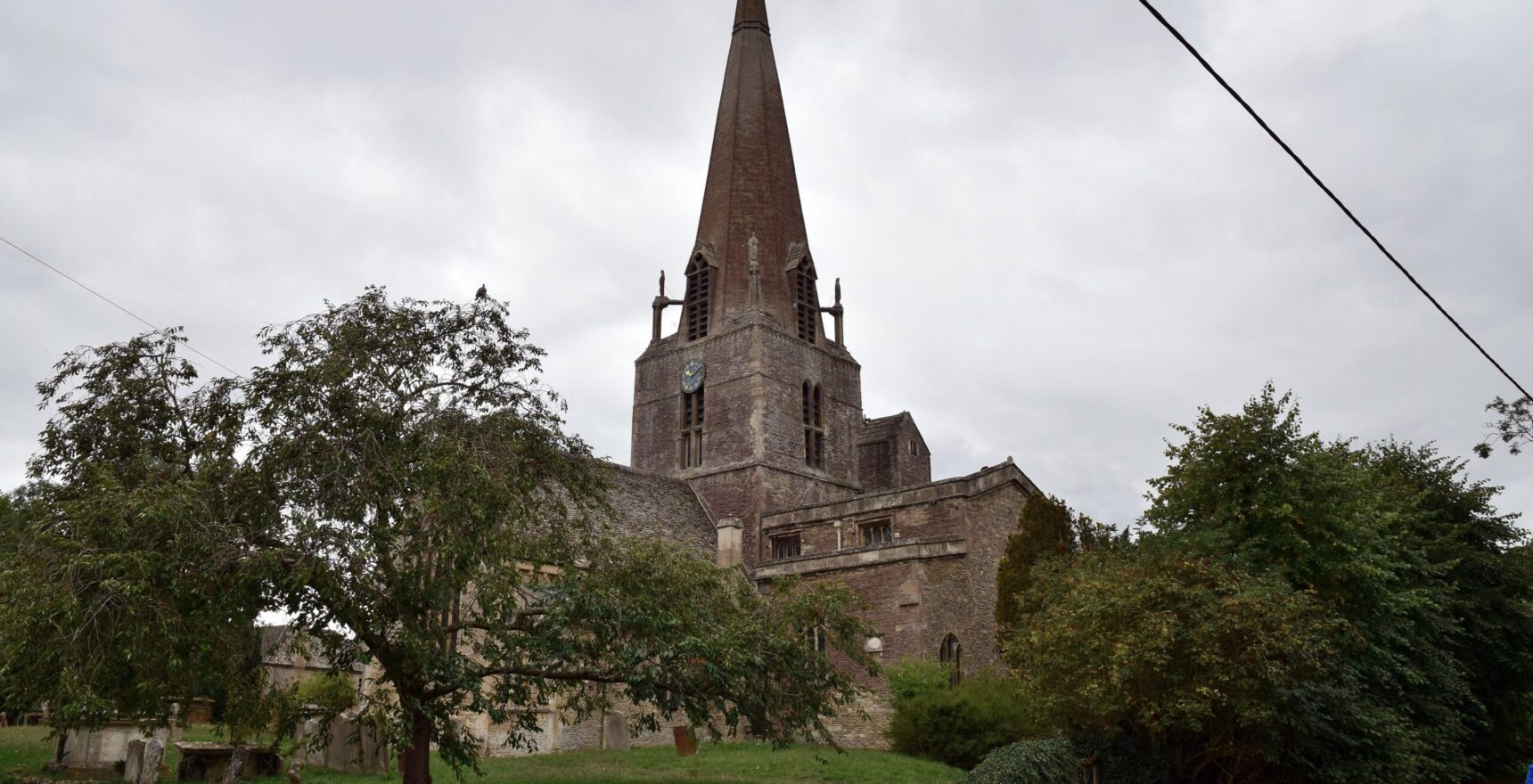 St Mary's Church in Bampton, Oxfordshire, with its tall stone spire and clock tower rising above the churchyard trees on an overcast day