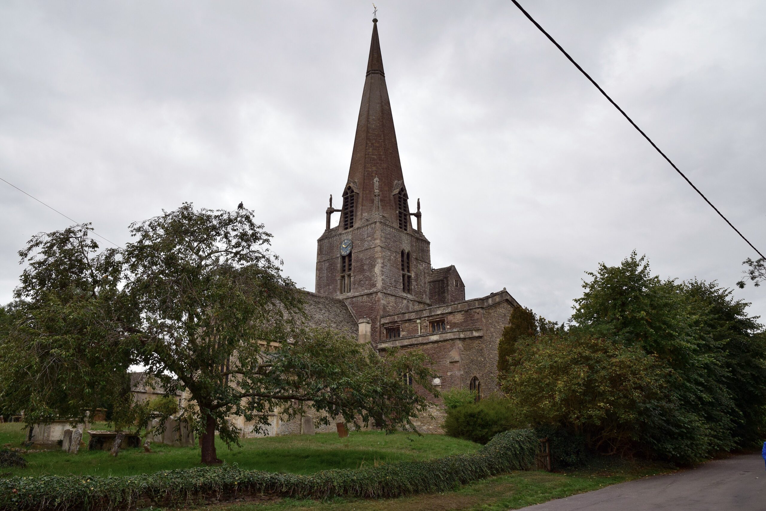 St Mary's Church in Bampton, Oxfordshire, with its tall stone spire and clock tower rising above the churchyard trees on an overcast day