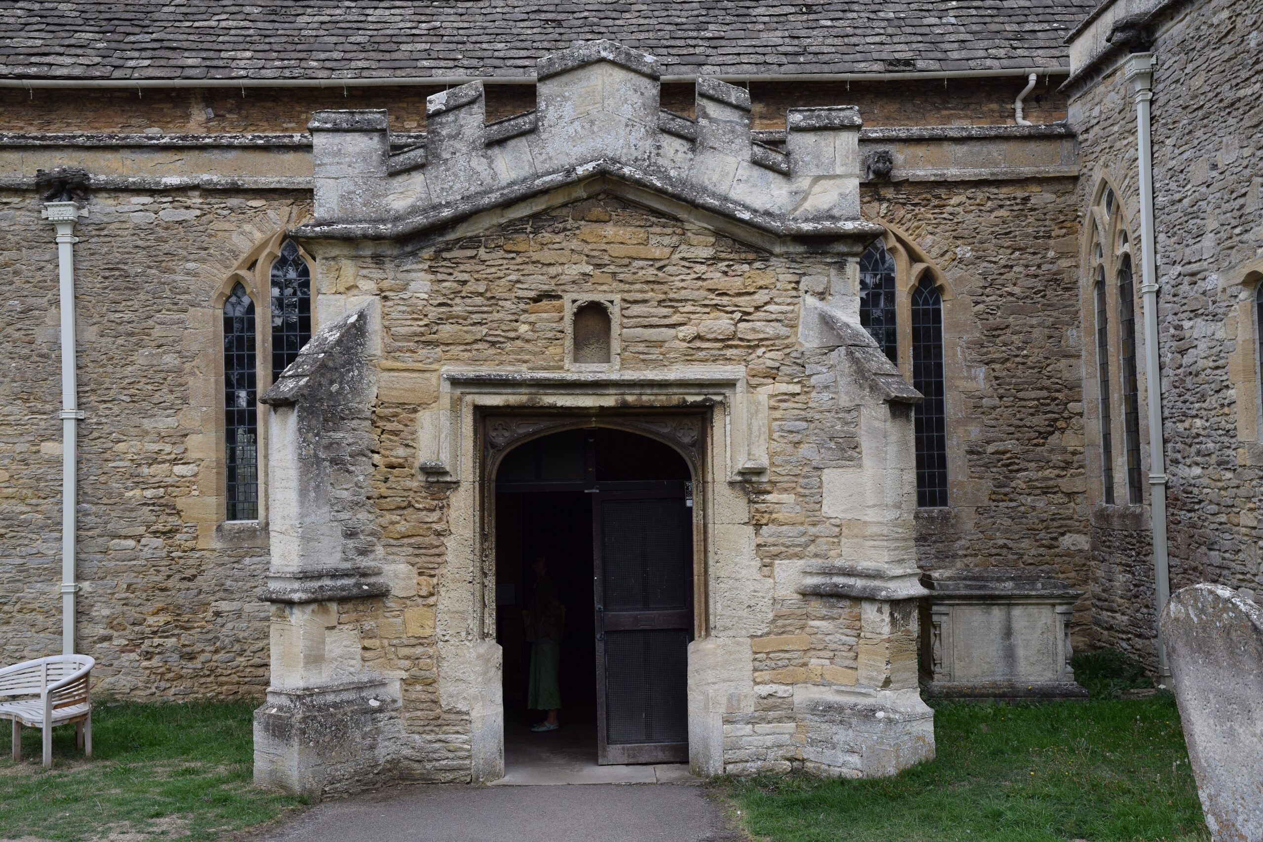 The battlemented stone porch of St Mary's Church in Bampton, with an open wooden door, a small empty niche above the doorway and lancet windows either side