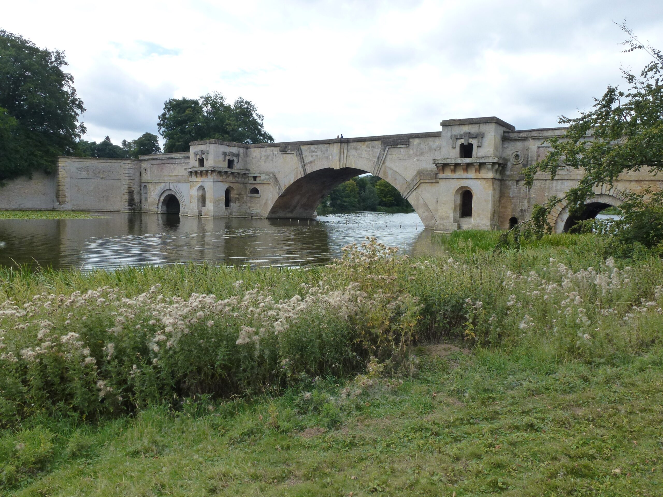 Vanbrugh's Grand Bridge at Blenheim Palace, a massive pale stone bridge with a great central arch and pavilion-like piers, crossing the lake behind a bank of thistles and long grass