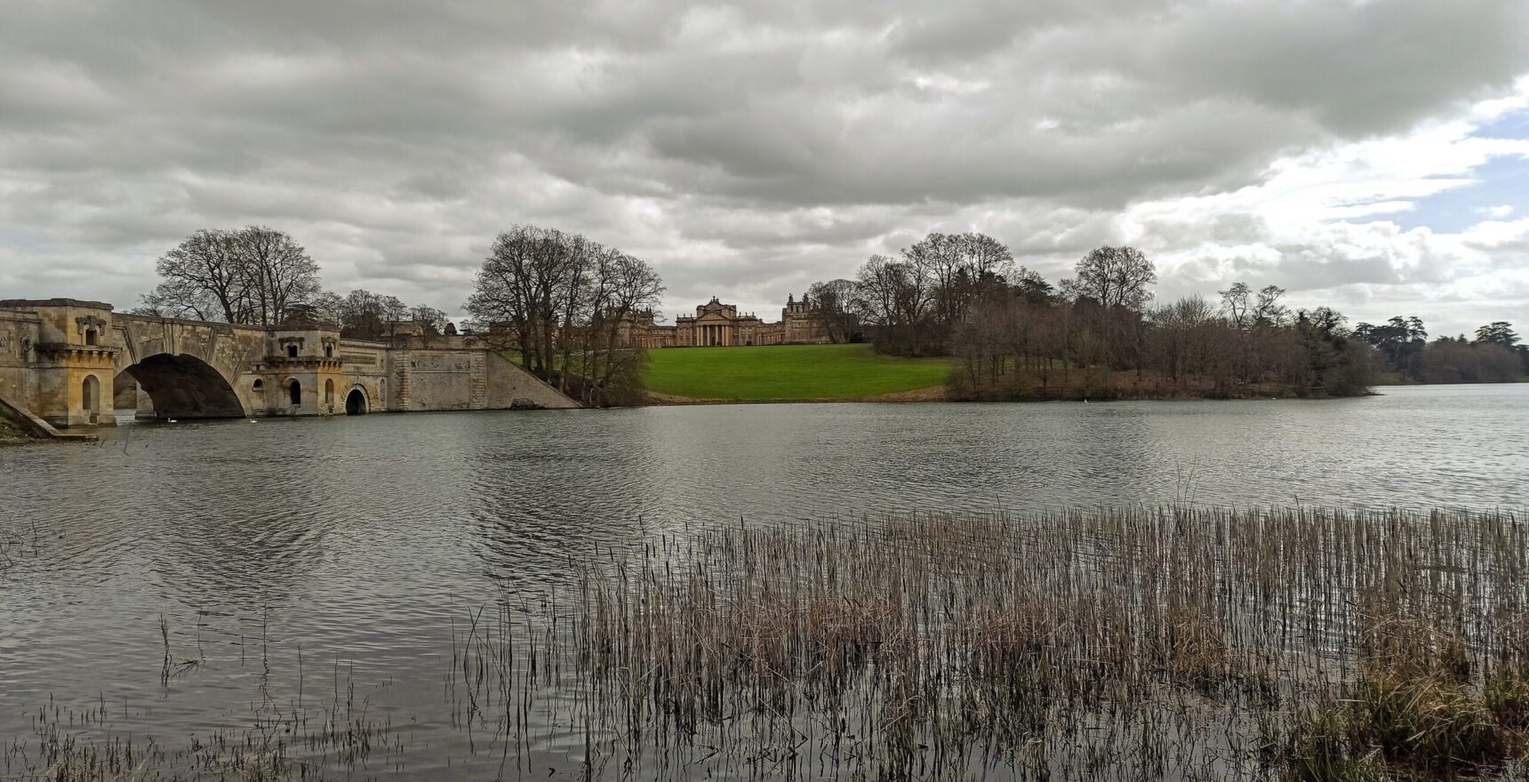 Blenheim Palace seen across the Great Lake under a heavy grey sky, with the arches of the Grand Bridge on the left, a green lawn rising to the palace and reeds in the foreground
