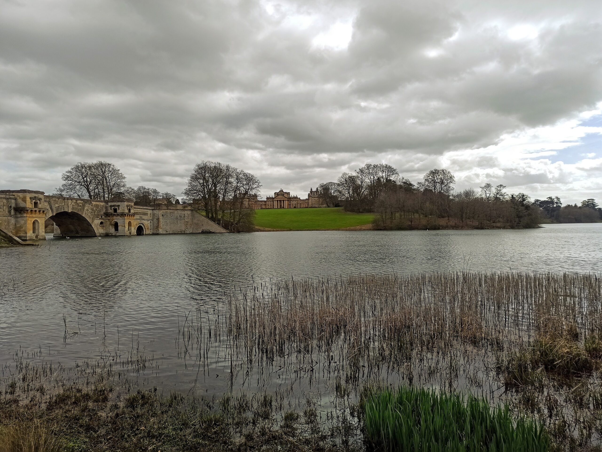 Blenheim Palace seen across the Great Lake under a heavy grey sky, with the arches of the Grand Bridge on the left, a green lawn rising to the palace and reeds in the foreground