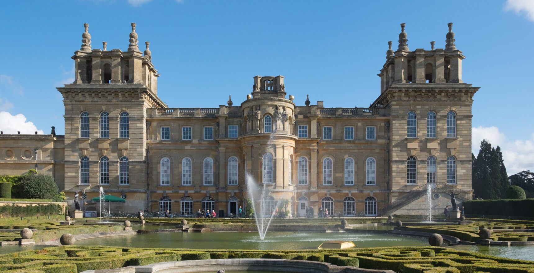 The west front of Blenheim Palace seen across the Water Terraces, with fountains playing in the pools and clipped box parterres under a blue sky