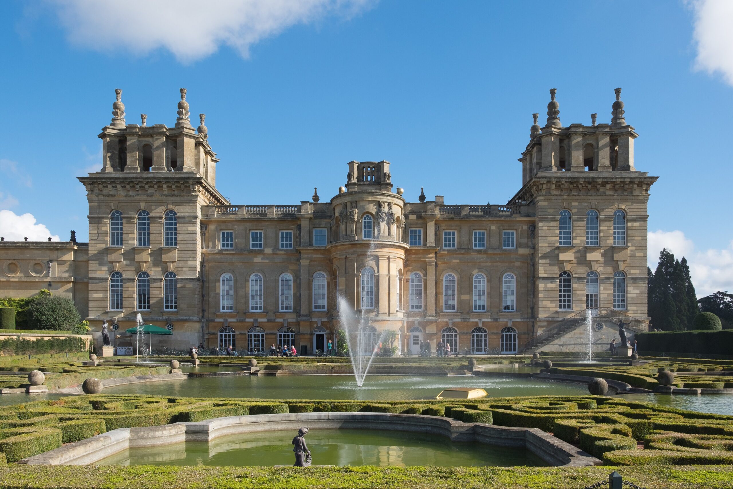 The west front of Blenheim Palace seen across the Water Terraces, with fountains playing in the pools and clipped box parterres under a blue sky