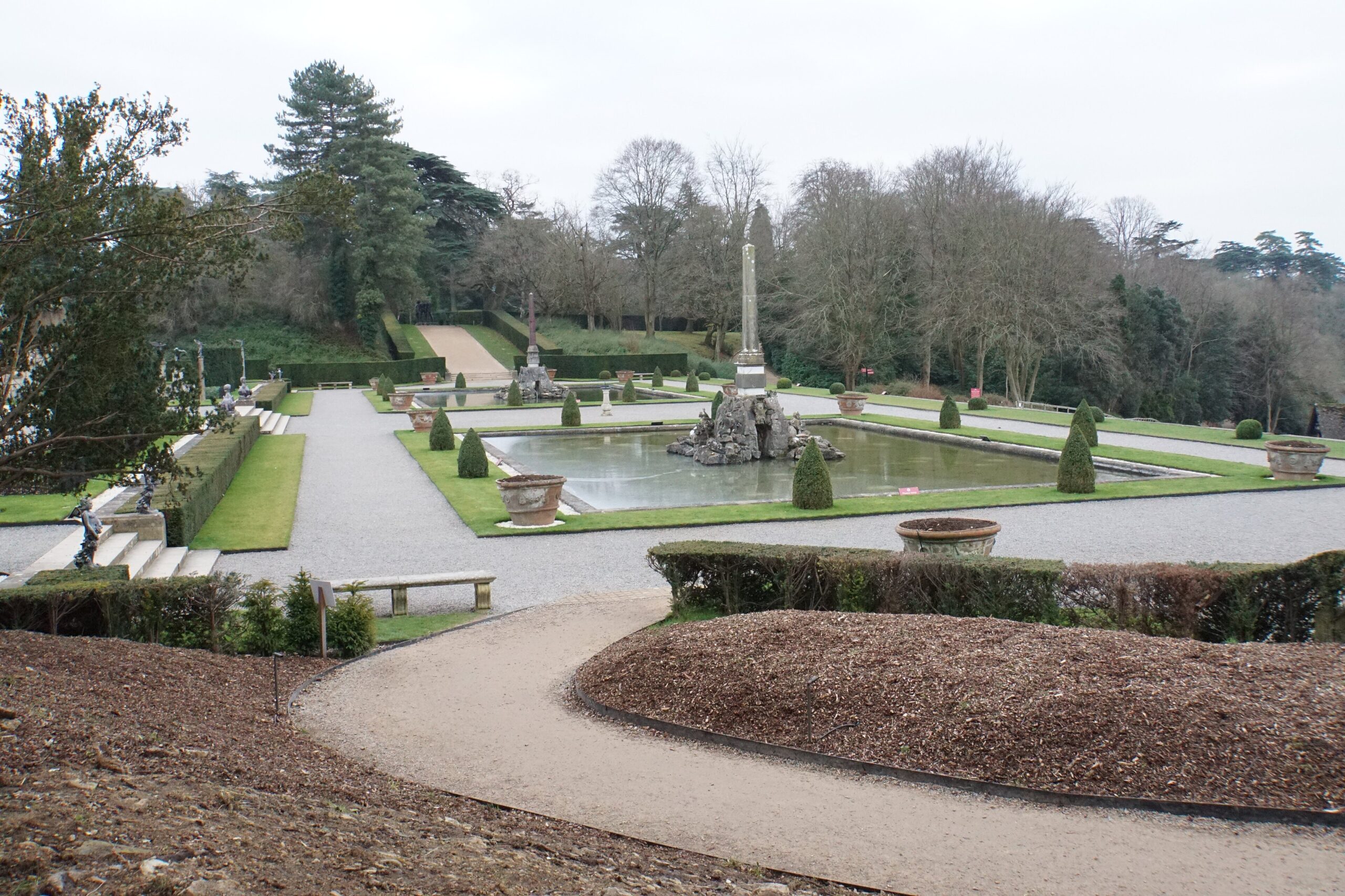 The formal Water Terraces at Blenheim Palace in winter, with rectangular pools, clipped yew cones, gravel walks and an obelisk fountain, and bare trees beyond