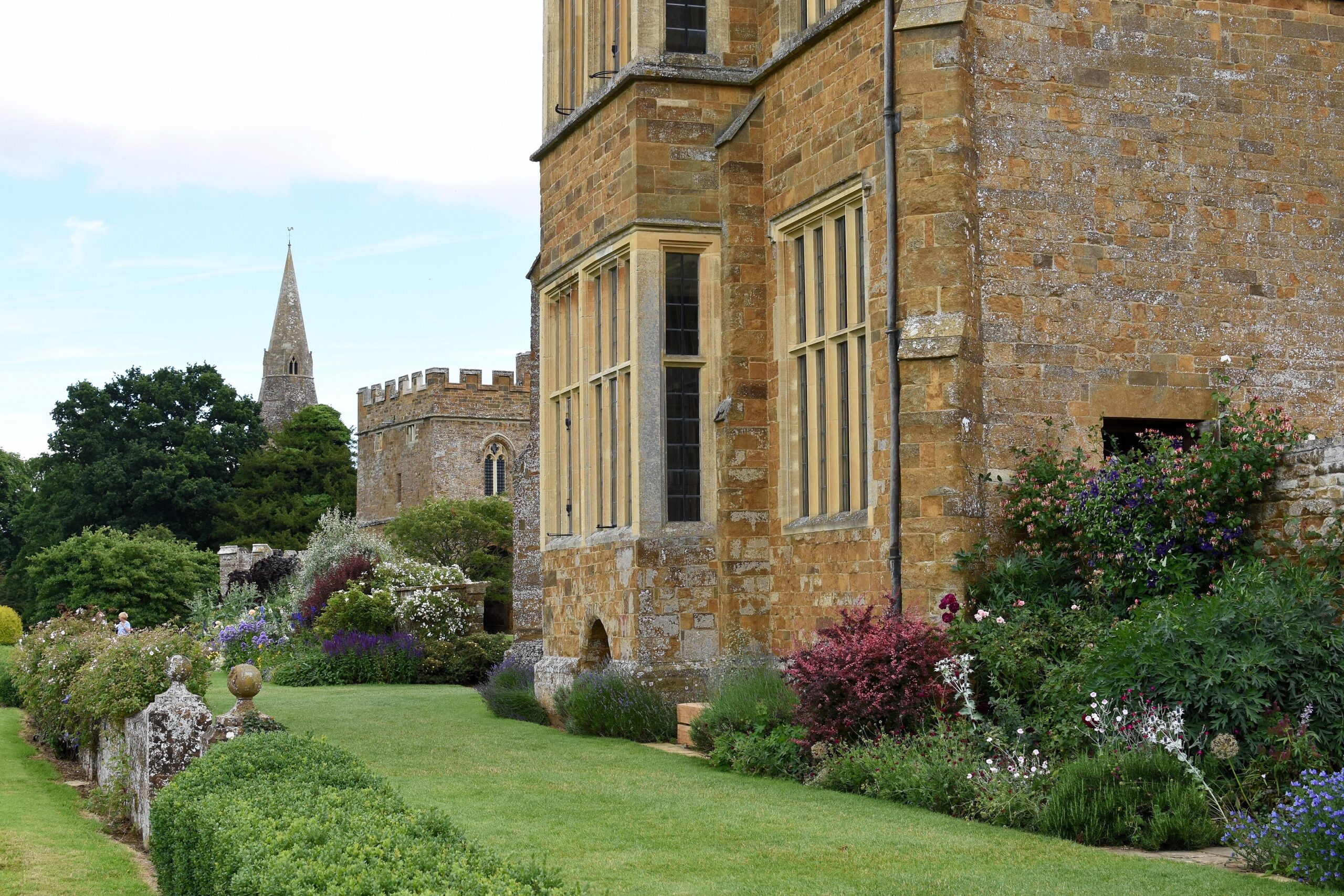 The honey-coloured stone walls and mullioned windows of Broughton Castle beside a flower-filled border and lawn, with a battlemented gatehouse and a church spire in the background