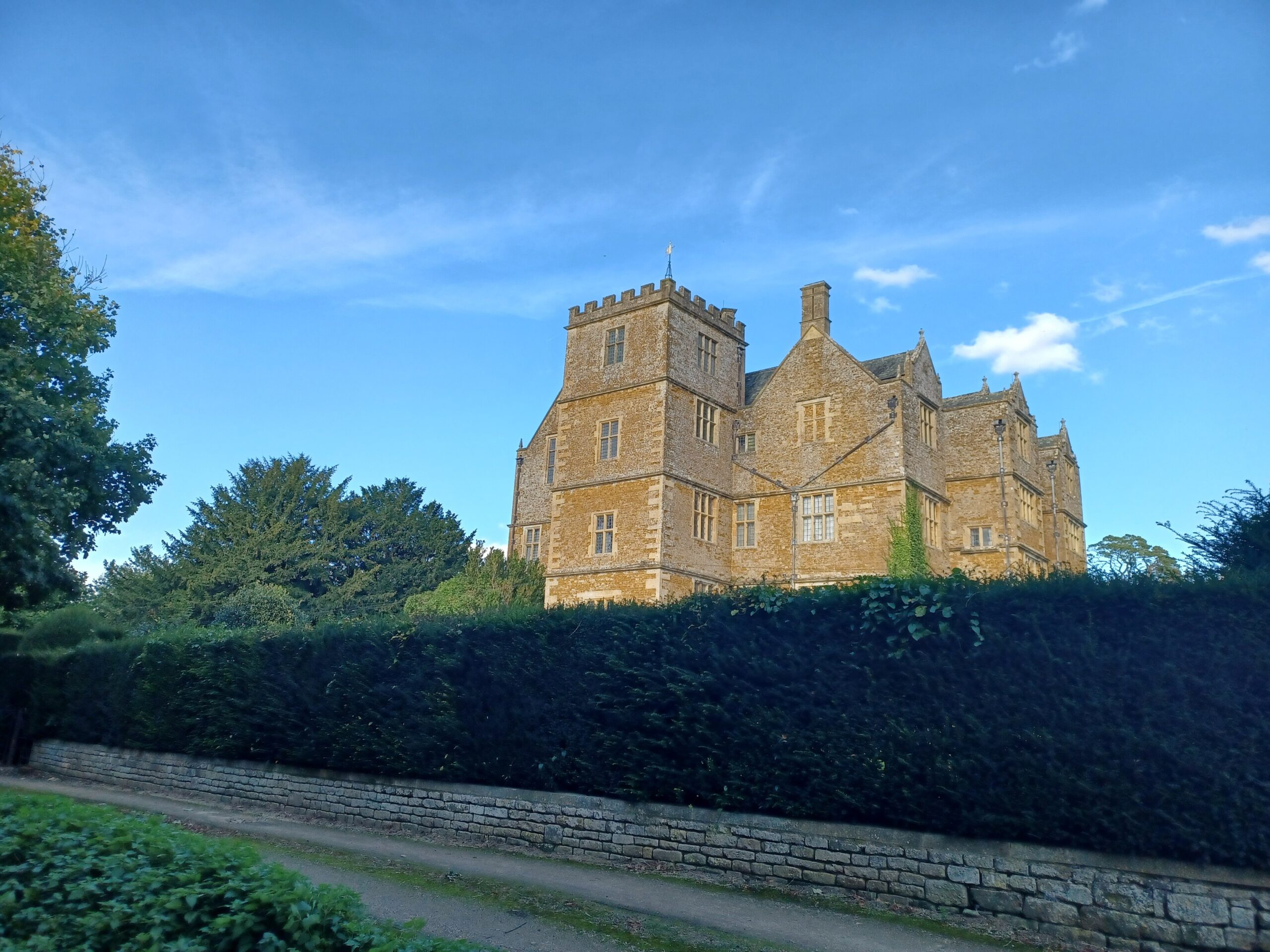 Chastleton House, a tall Jacobean manor of golden stone with a battlemented tower and gabled wings, rising above a clipped yew hedge and a dry-stone wall under a blue sky