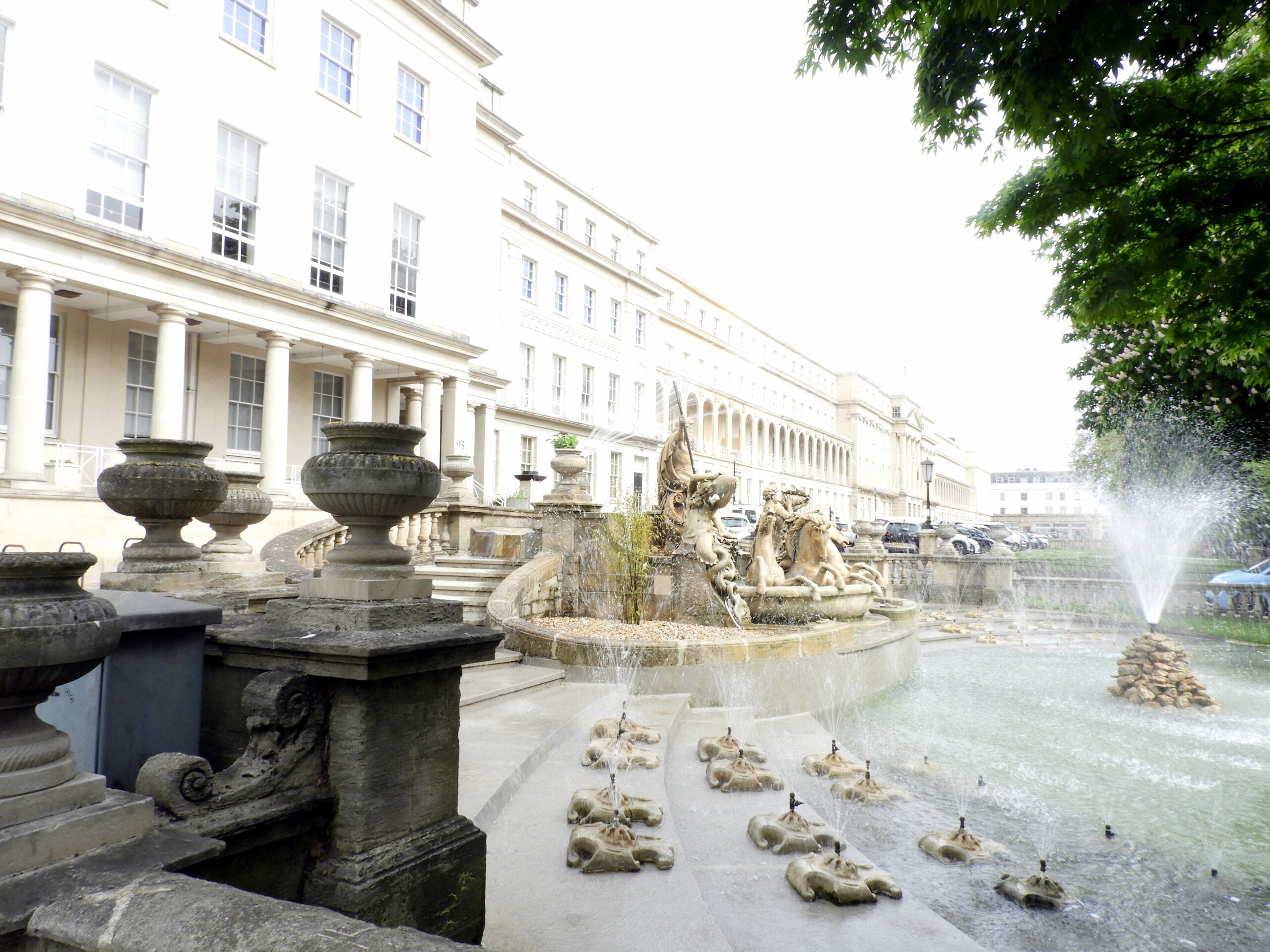 Neptune's Fountain in Cheltenham with water jets in front of a long Regency terrace with stone urns and a colonnade