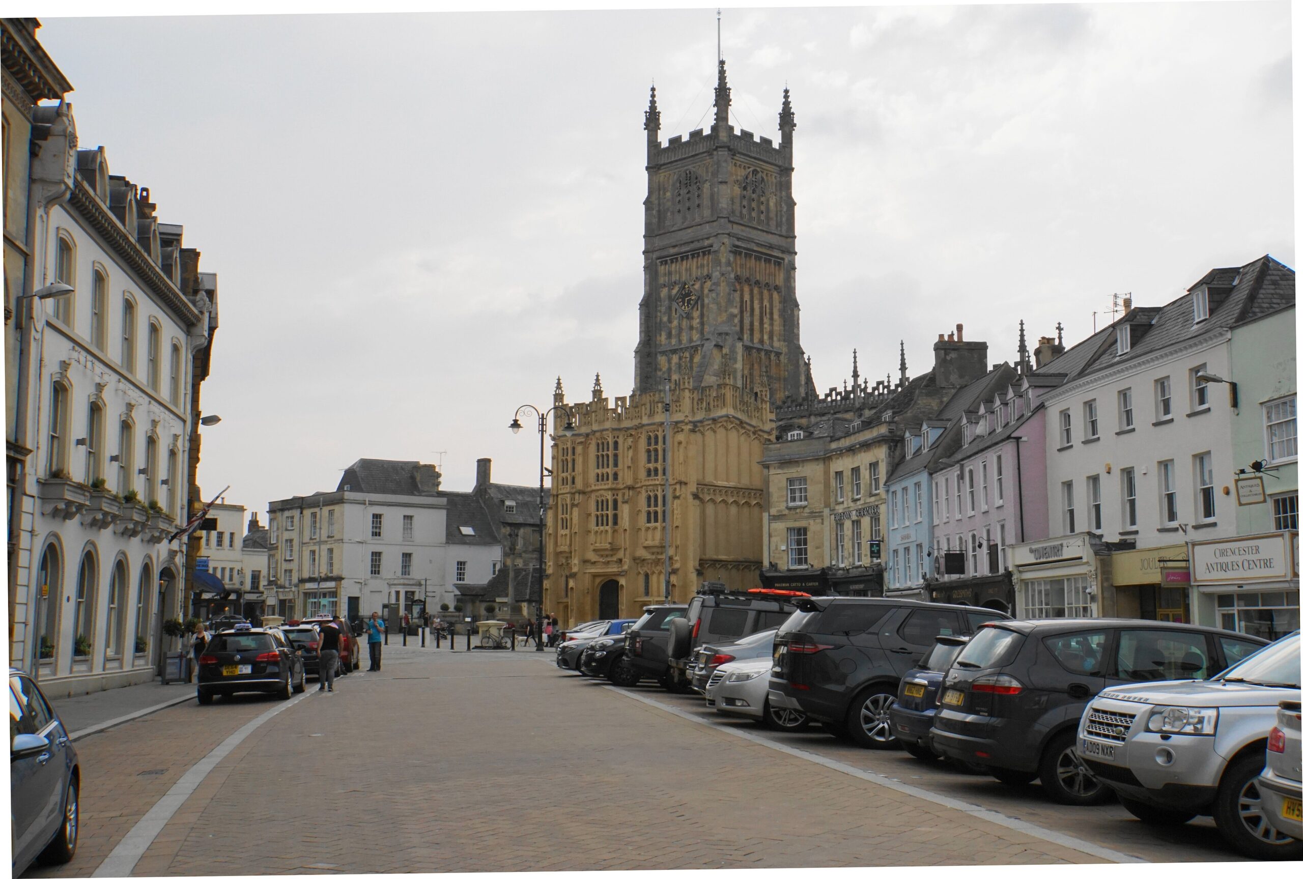 Cirencester Market Place with parked cars, shopfronts and the tall Perpendicular tower and porch of St John Baptist church