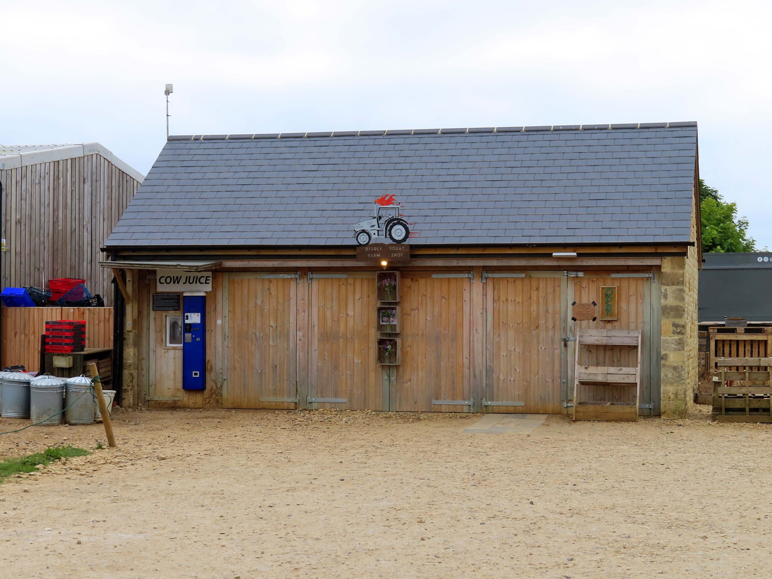 The timber-clad Diddly Squat Farm Shop barn with a stone gable, a tractor-and-flames sign on the roof and a blue Cow Juice milk vending machine by the door