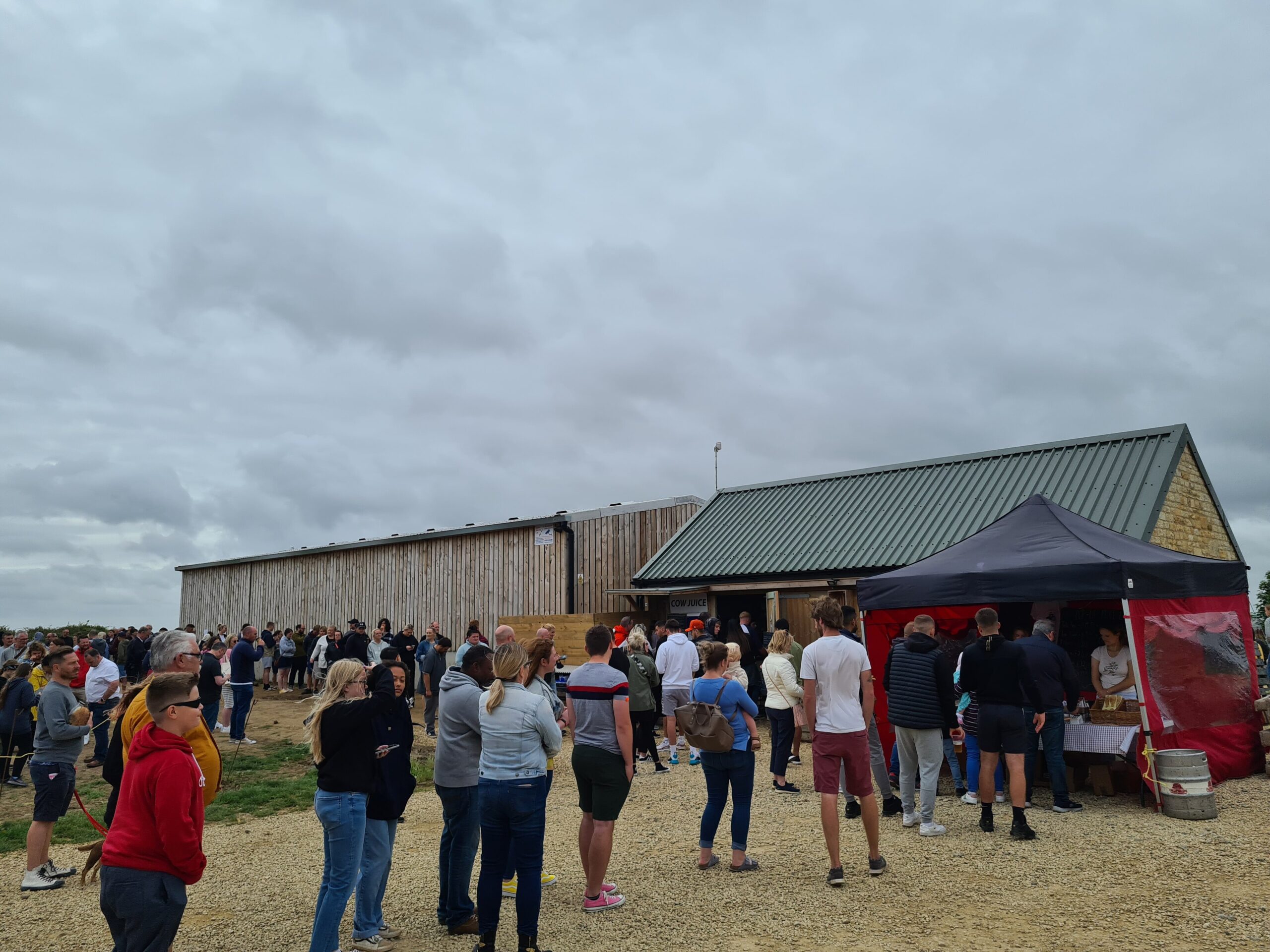 A long queue of visitors on the gravel yard outside Diddly Squat Farm Shop under a grey sky, with a red and black drinks gazebo beside the barn