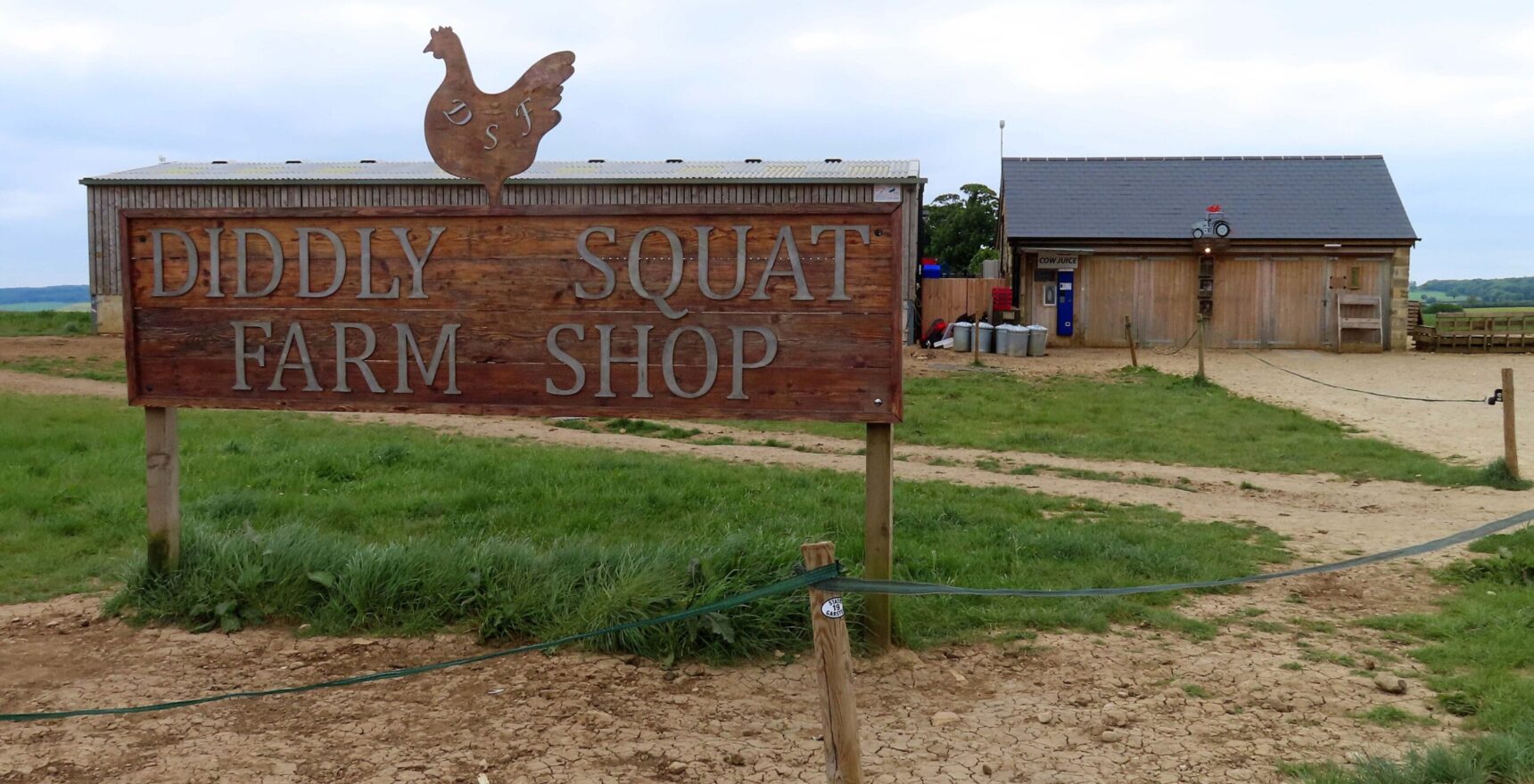 The large wooden Diddly Squat Farm Shop sign topped with a metal hen, with the timber and stone shop barn behind it on the farm near Chadlington