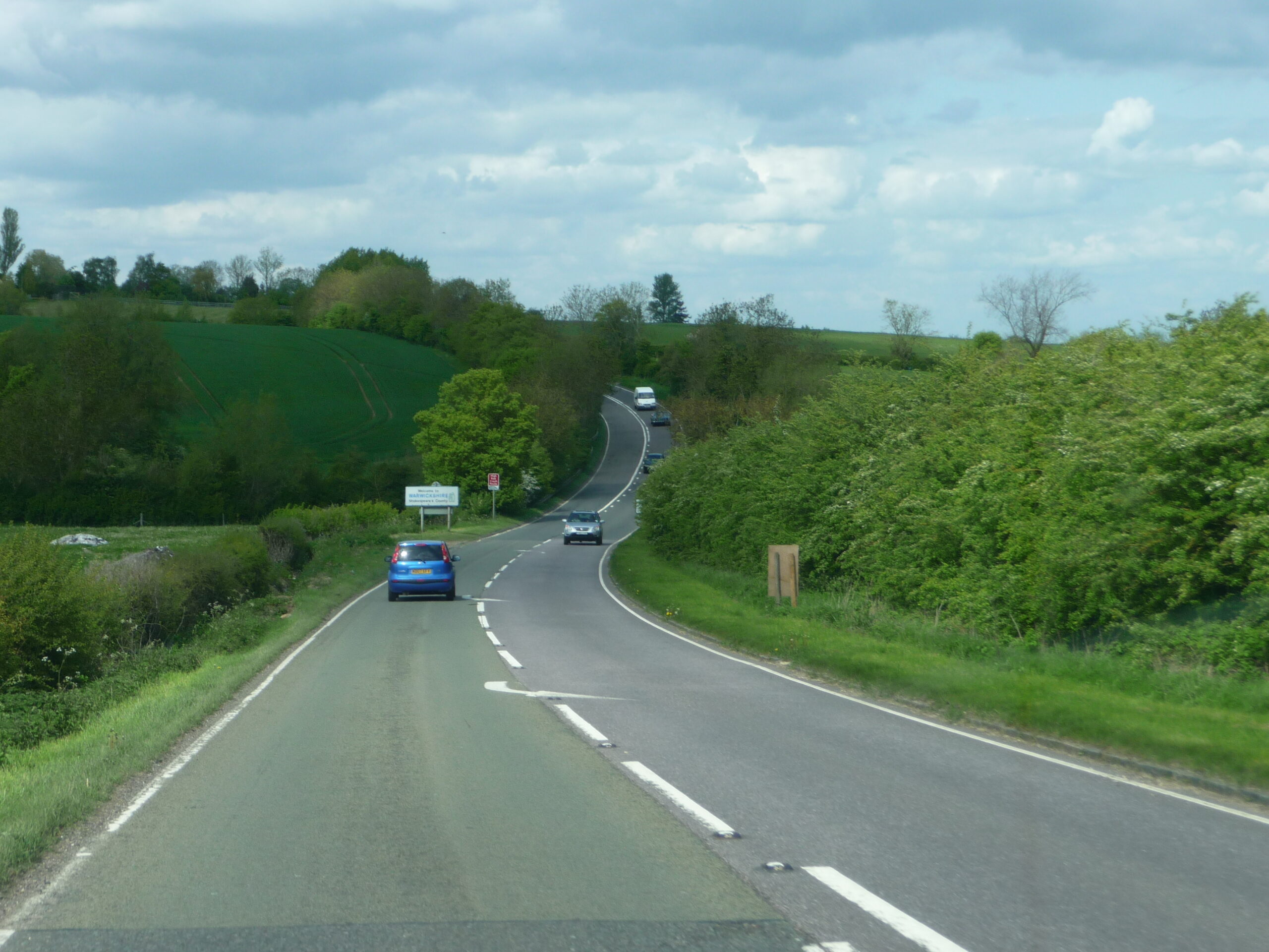 The Fosse Way winding over rolling green countryside between hedgerows, with cars on the road and a Welcome to Warwickshire sign by the verge