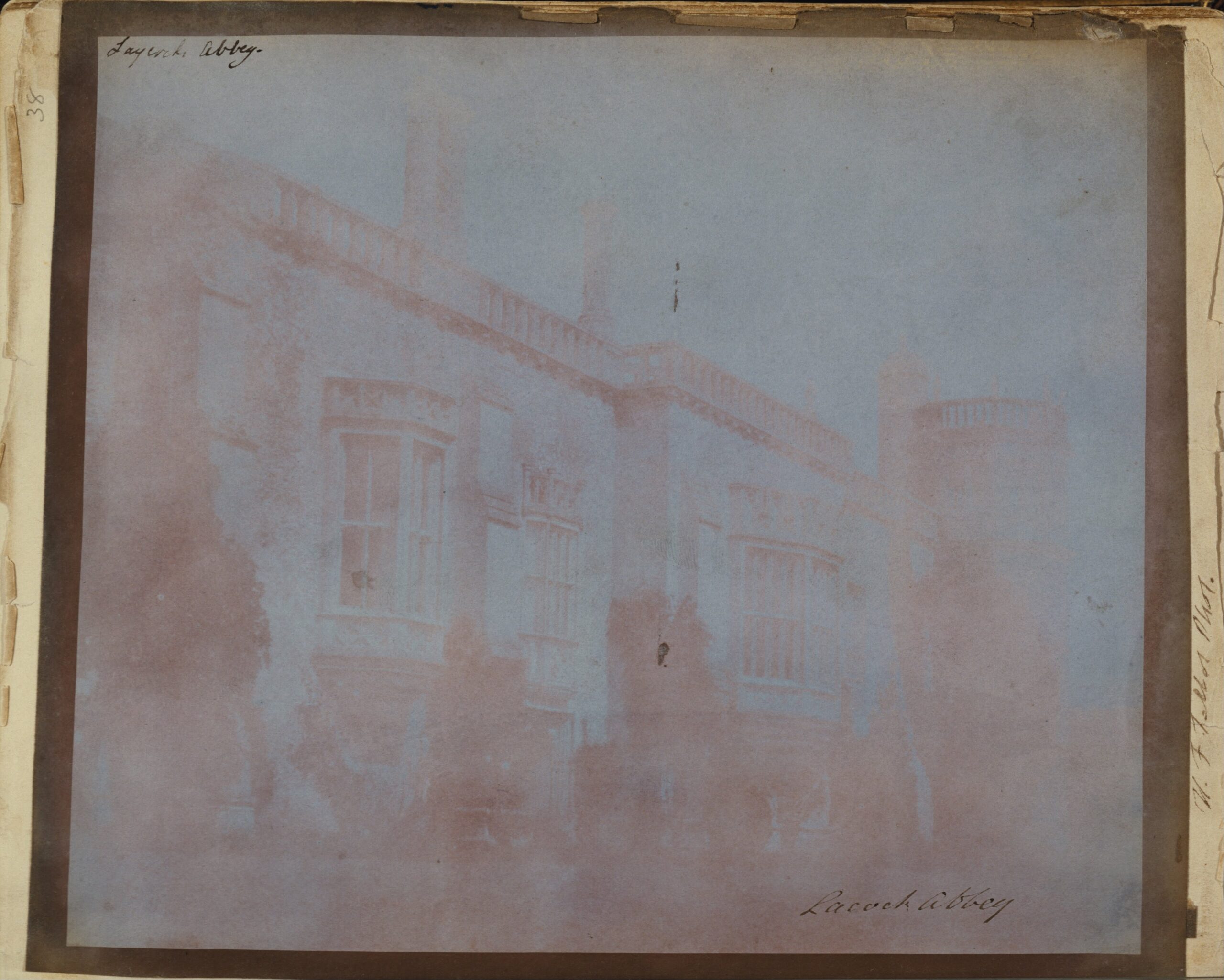 A faded early salted-paper photograph by William Henry Fox Talbot of the south front of Lacock Abbey, showing oriel windows and a balustraded roofline, with the words Lacock Abbey handwritten on the mount