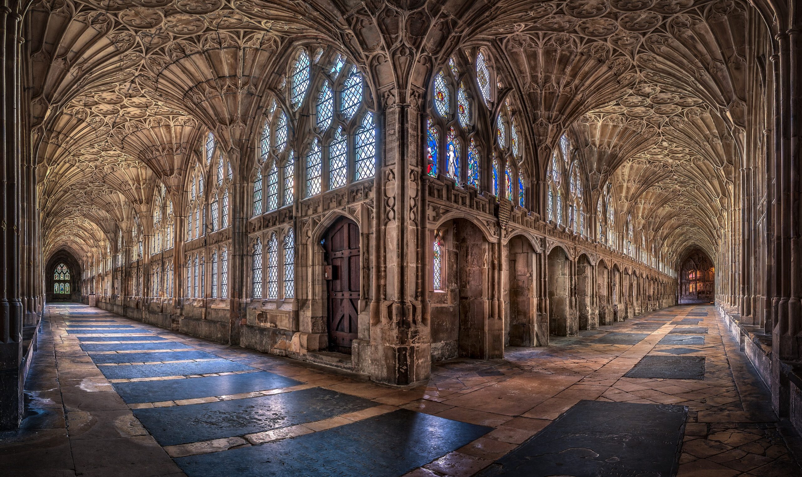 A corner of the Gloucester Cathedral cloisters where two fan-vaulted walks meet, with tall leaded windows, a wooden door and worn flagstones stretching away in both directions
