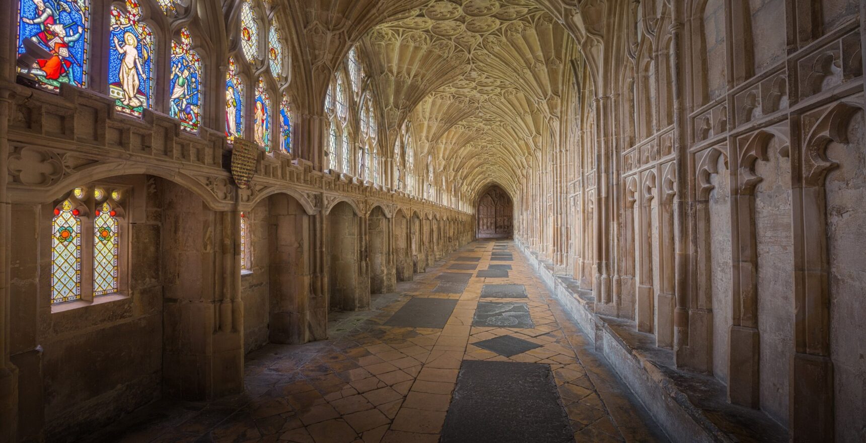 The fan-vaulted south walk of the Gloucester Cathedral cloisters, with stained-glass windows on the left and worn flagstones running into the distance