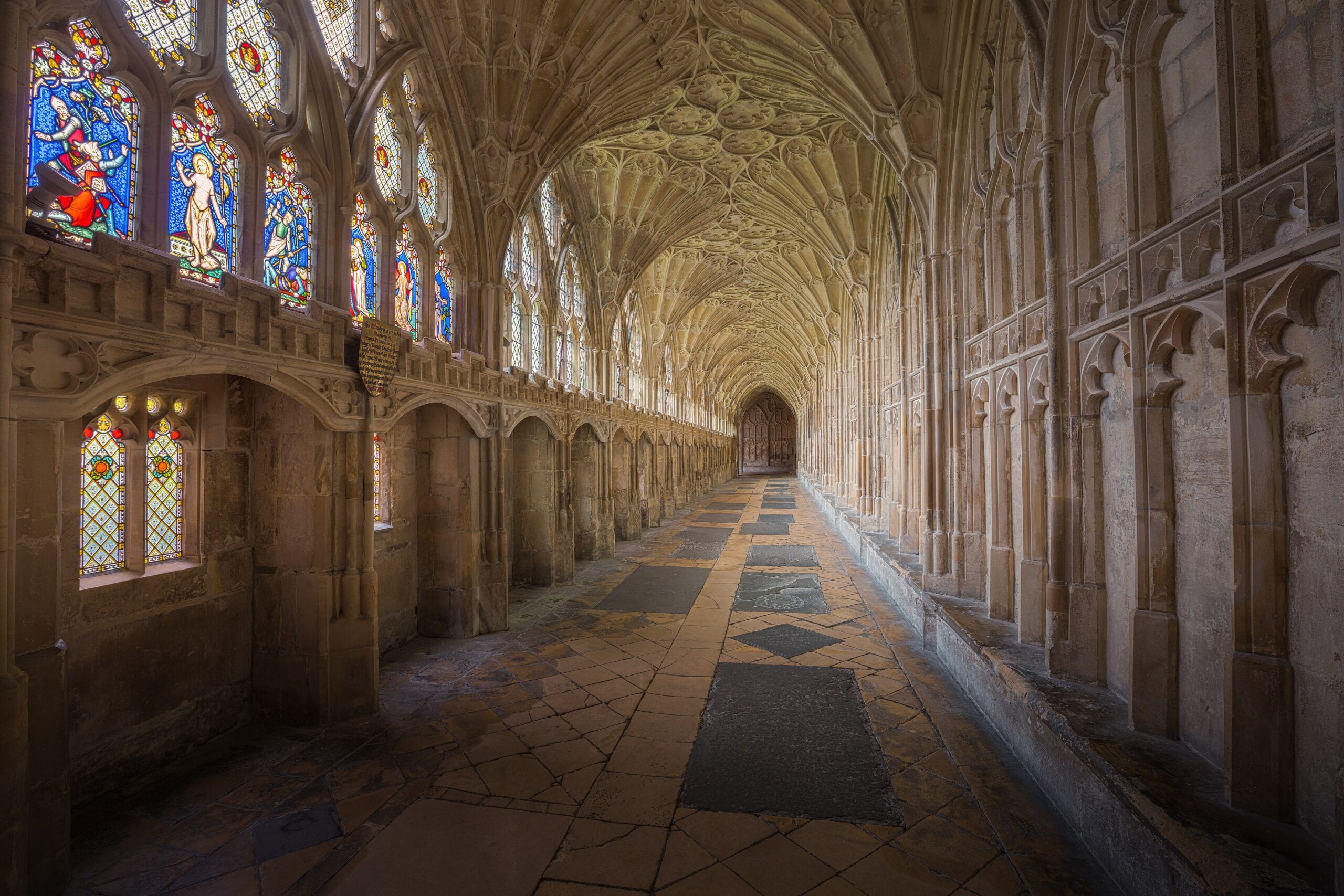 The fan-vaulted south walk of the Gloucester Cathedral cloisters, with stained-glass windows on the left and worn flagstones running into the distance