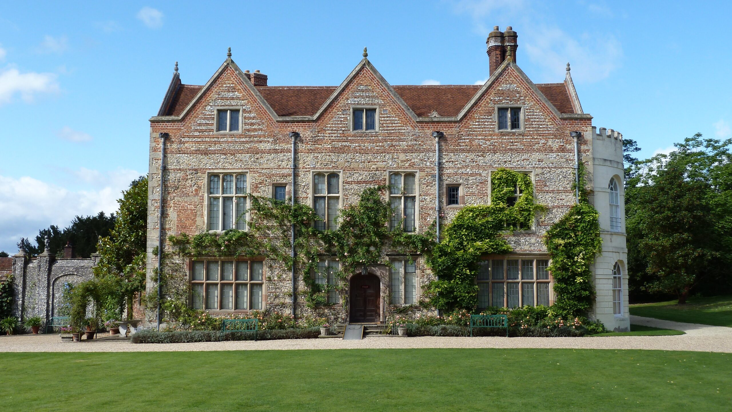 The brick and flint Tudor front of Greys Court near Henley-on-Thames, with three gables, climbing roses and a manicured lawn in sunshine