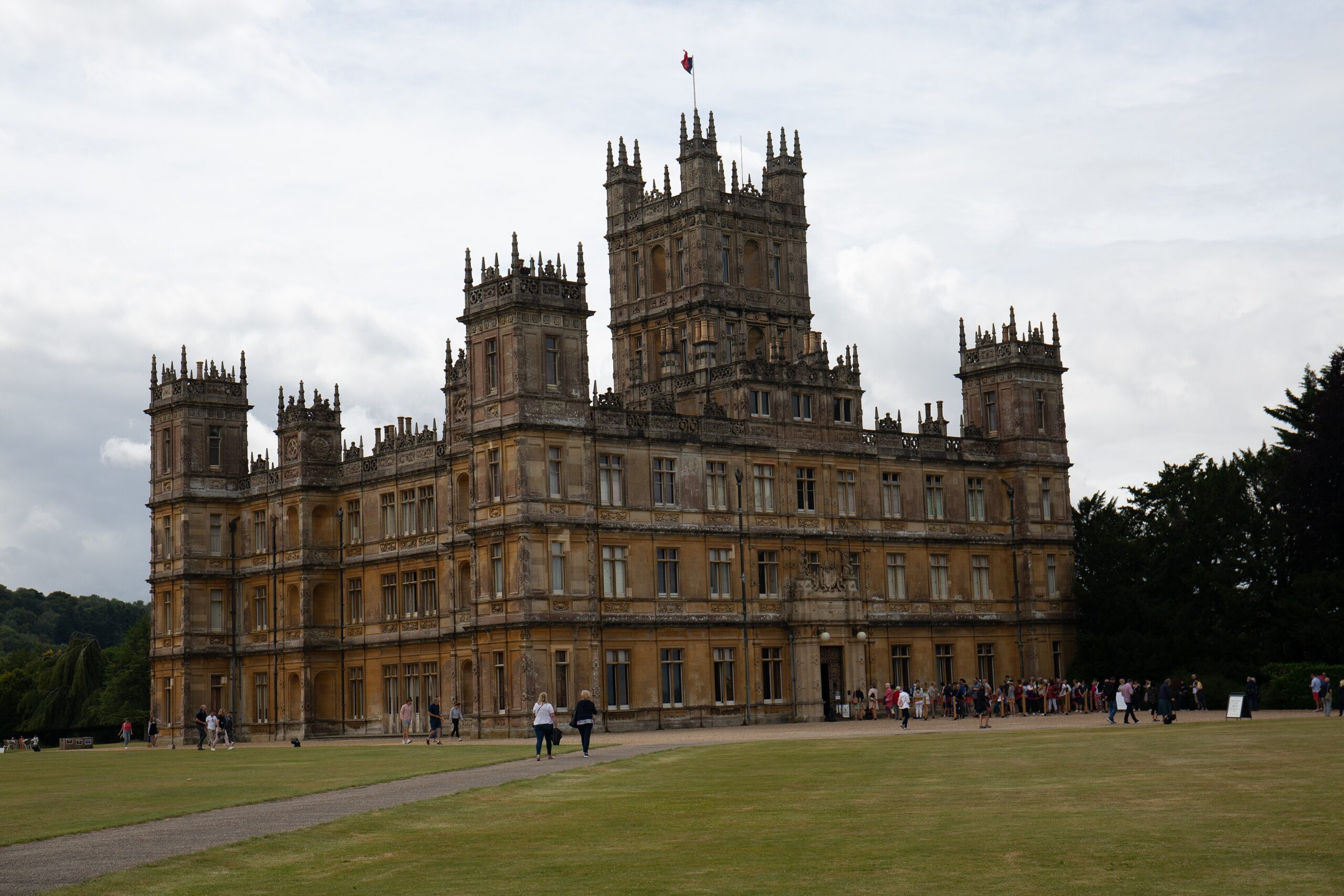 The pinnacled golden-stone front of Highclere Castle under a cloudy sky, with visitors walking on the gravel drive and queuing at the entrance across a wide lawn