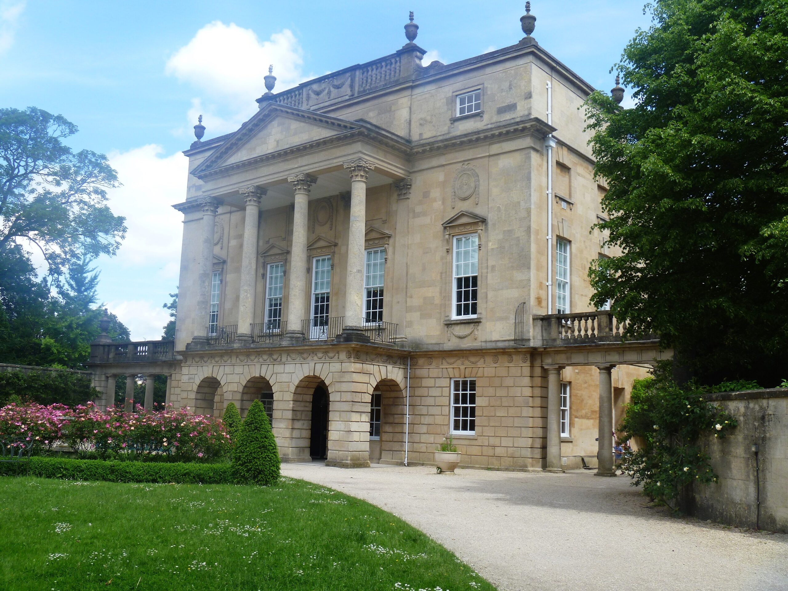 The Holburne Museum in Bath, a pale stone Georgian building with a columned portico, balustrade and urns, behind a lawn and pink roses