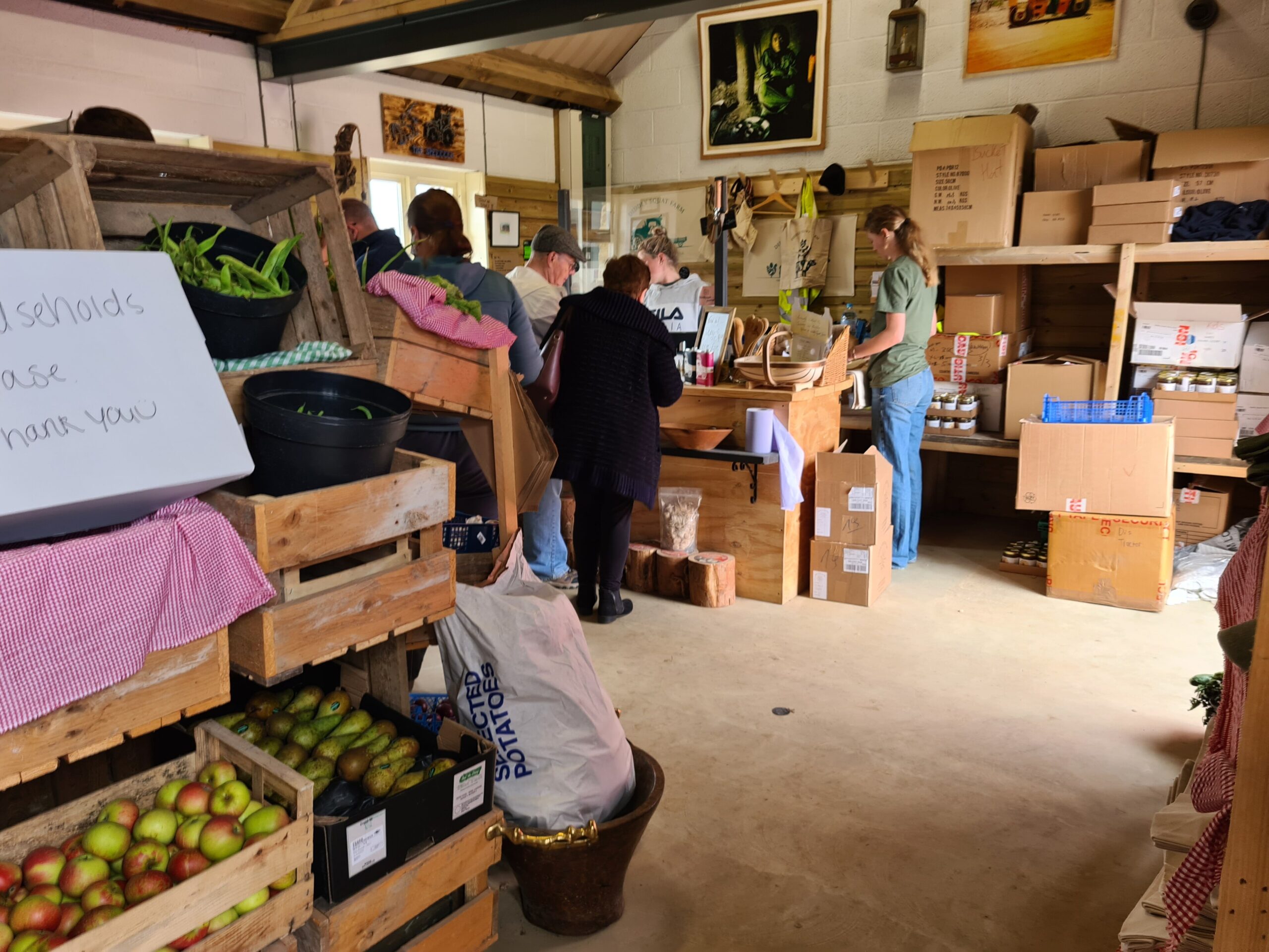 Inside the Diddly Squat Farm Shop, with wooden crates of apples and pears, shoppers at the counter, tote bags and caps on hooks and stacked boxes of stock