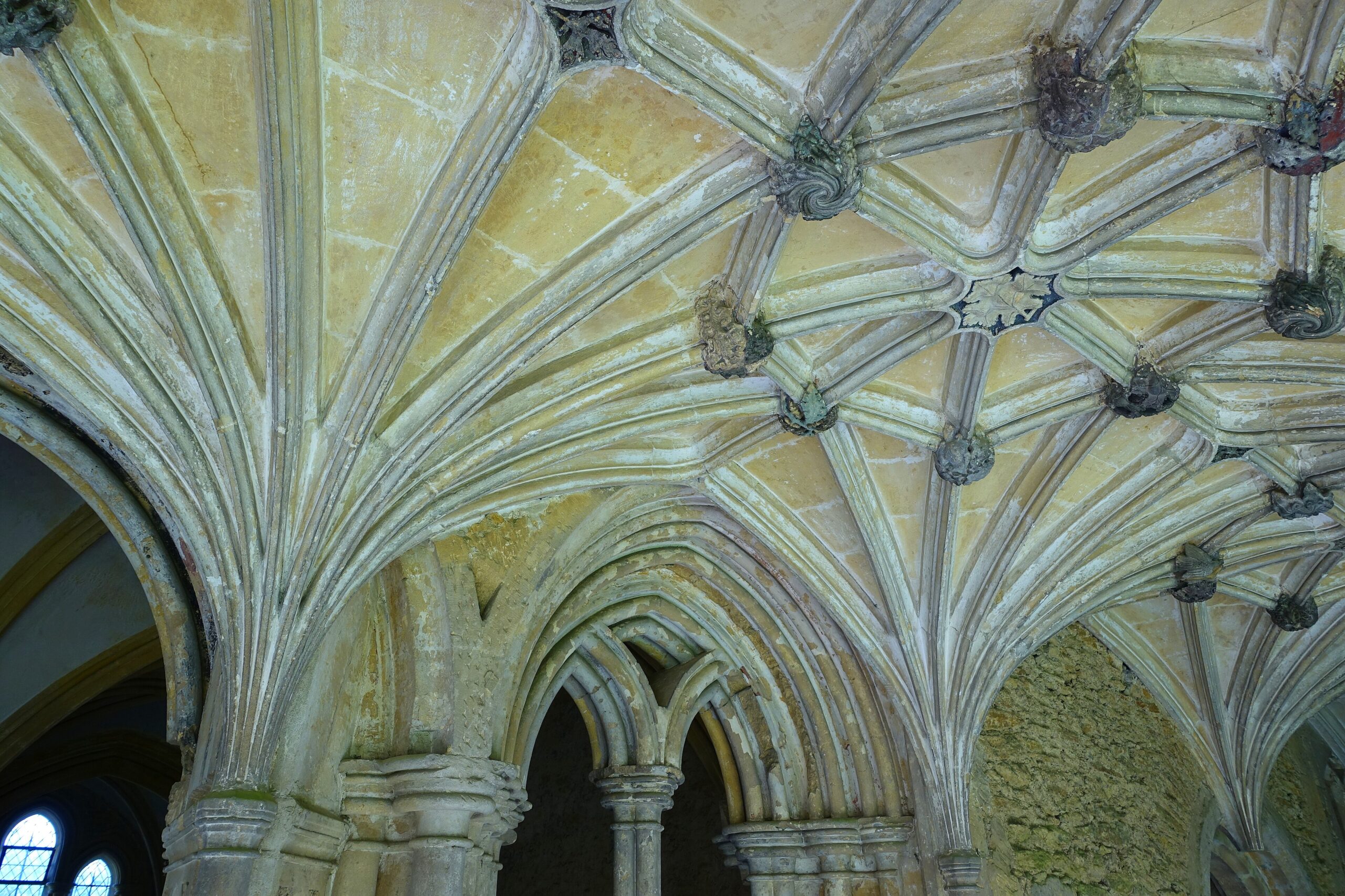 Pale stone ribbed vaulting in the cloisters of Lacock Abbey, with carved bosses at the junctions of the ribs and pointed Gothic arches below