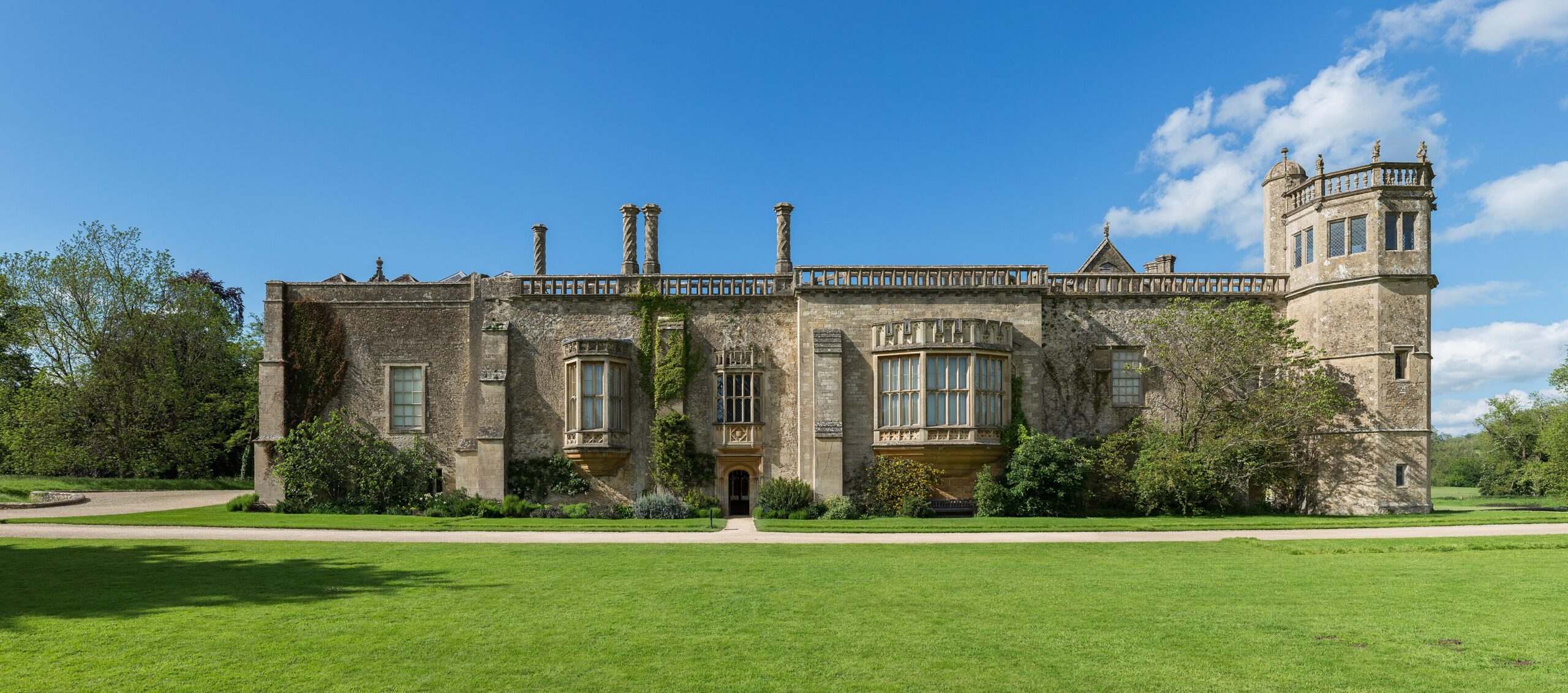 The long south front of Lacock Abbey in Wiltshire, with oriel windows, a balustraded roofline, twisted chimneys and an octagonal tower, above a wide lawn under a blue sky