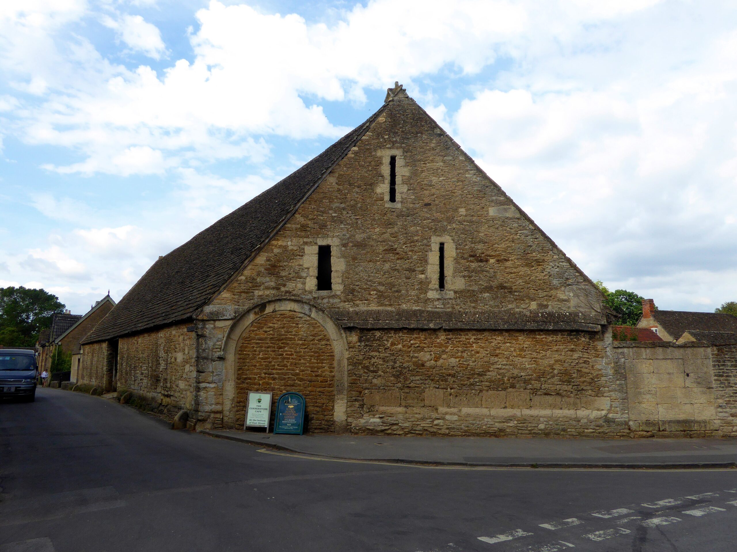 The medieval stone tithe barn in Lacock with a steep stone-tiled roof, narrow slit windows and a large blocked archway, at a street corner under a cloudy sky