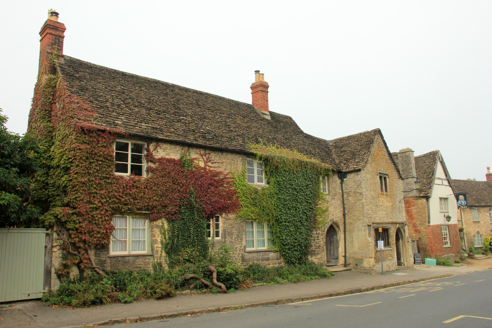 Old stone houses in Lacock with stone-tiled roofs and tall red-brick chimneys, their walls covered in Virginia creeper turning red and green, beside a quiet village street