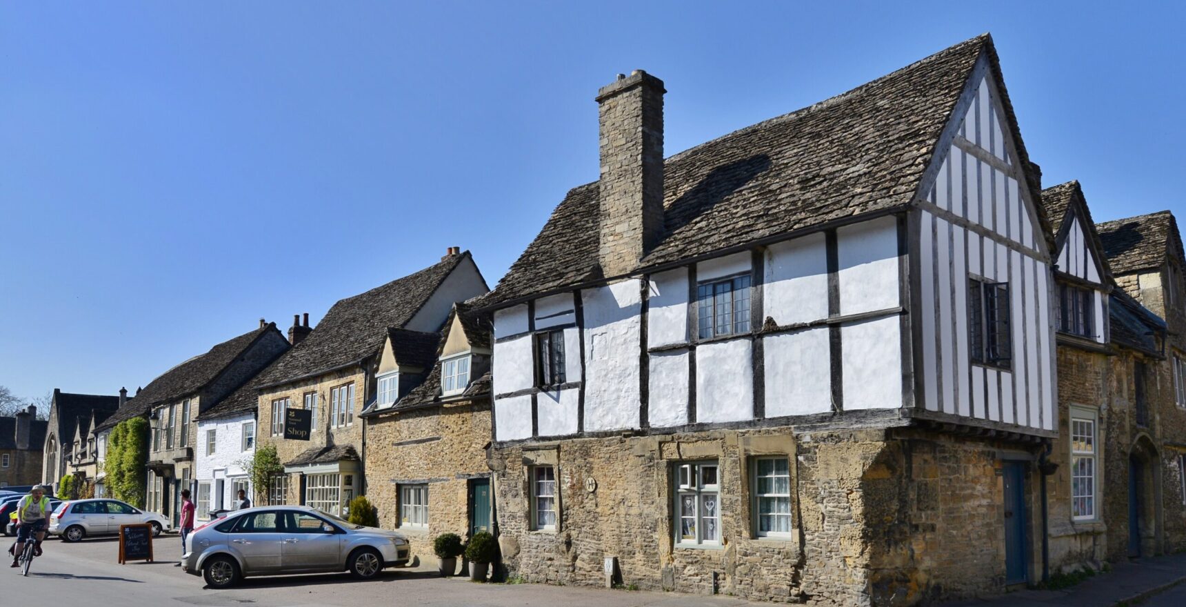The corner of East Street and the High Street in Lacock, with a jettied timber-framed house on a stone ground floor and a row of stone houses and the National Trust shop beyond