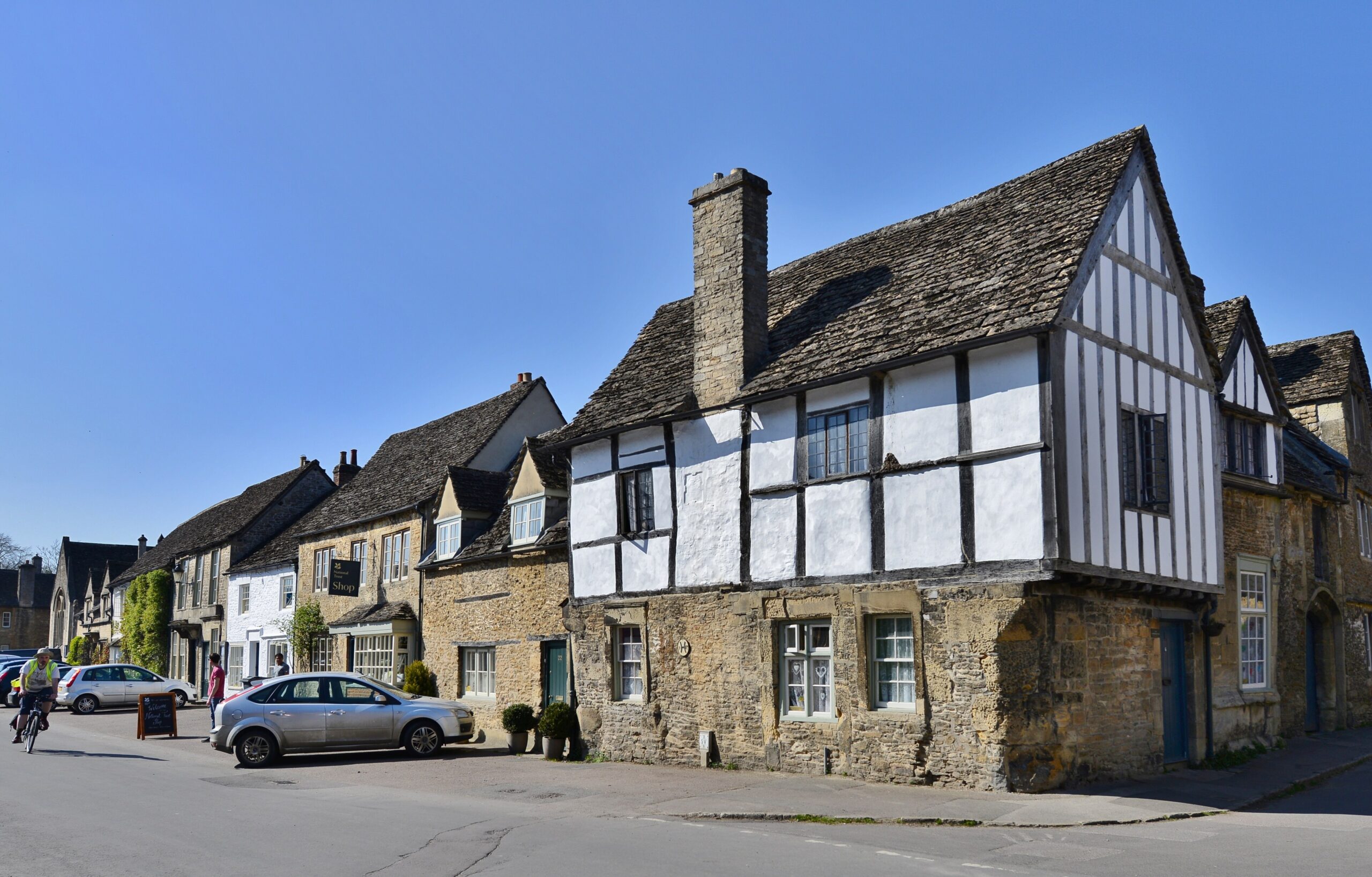 The corner of East Street and the High Street in Lacock, with a jettied timber-framed house on a stone ground floor and a row of stone houses and the National Trust shop beyond