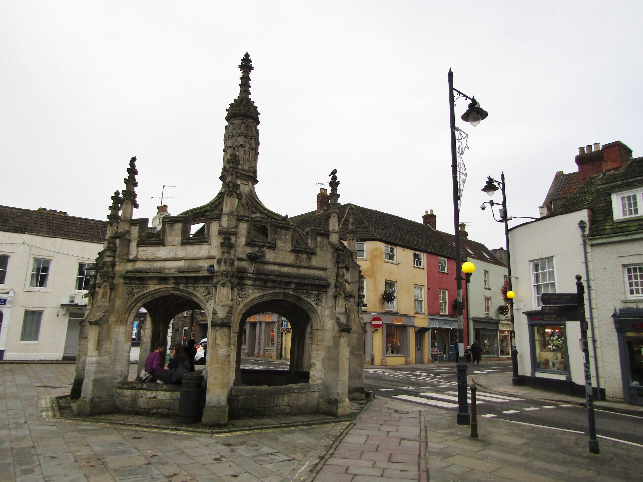 The octagonal late-medieval Market Cross at Malmesbury with pinnacles and open arches, with people sitting beneath it