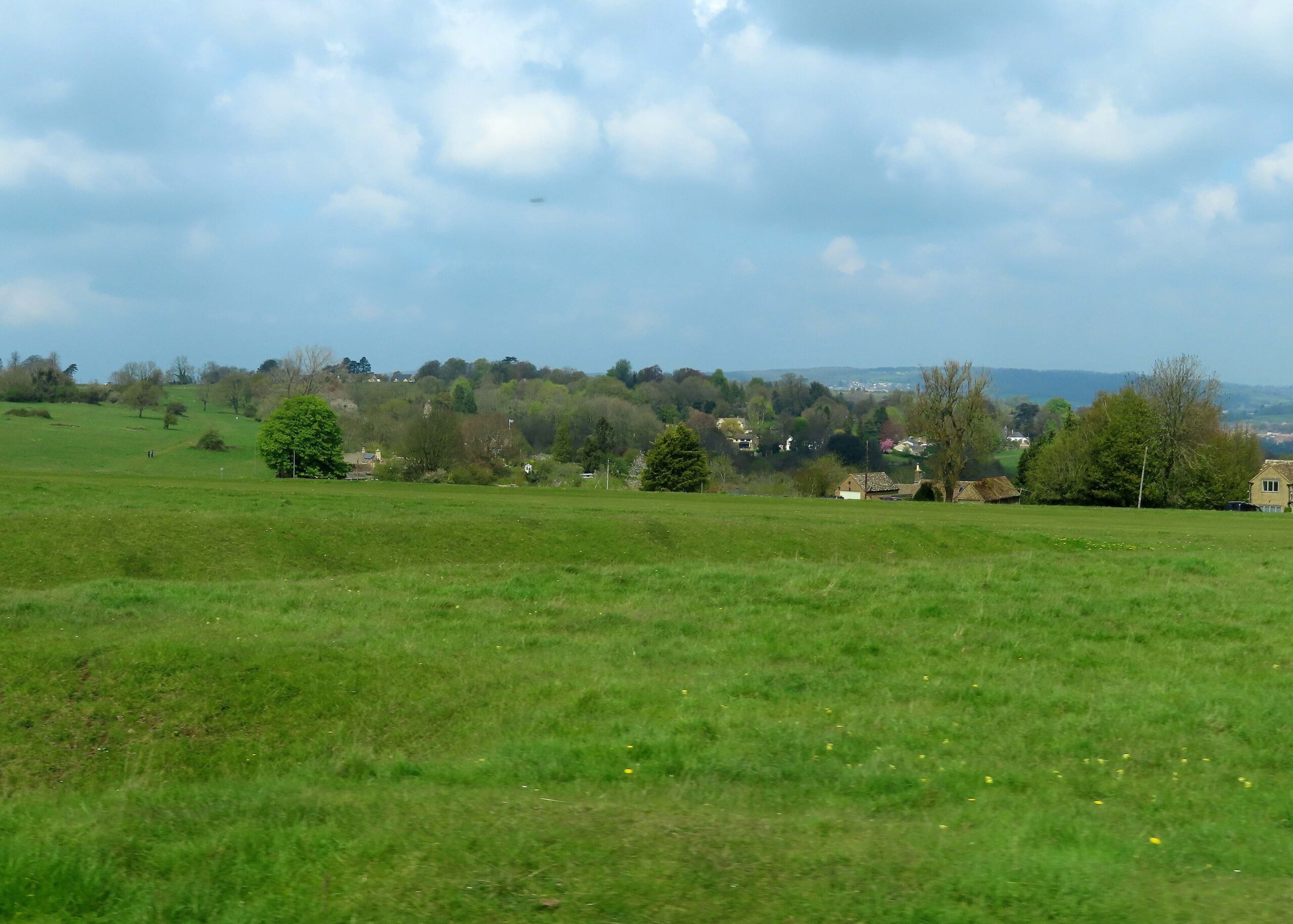 Low grassy banks and ditches of the Bulwarks earthworks running across Minchinhampton Common, with trees and stone houses at the common's edge