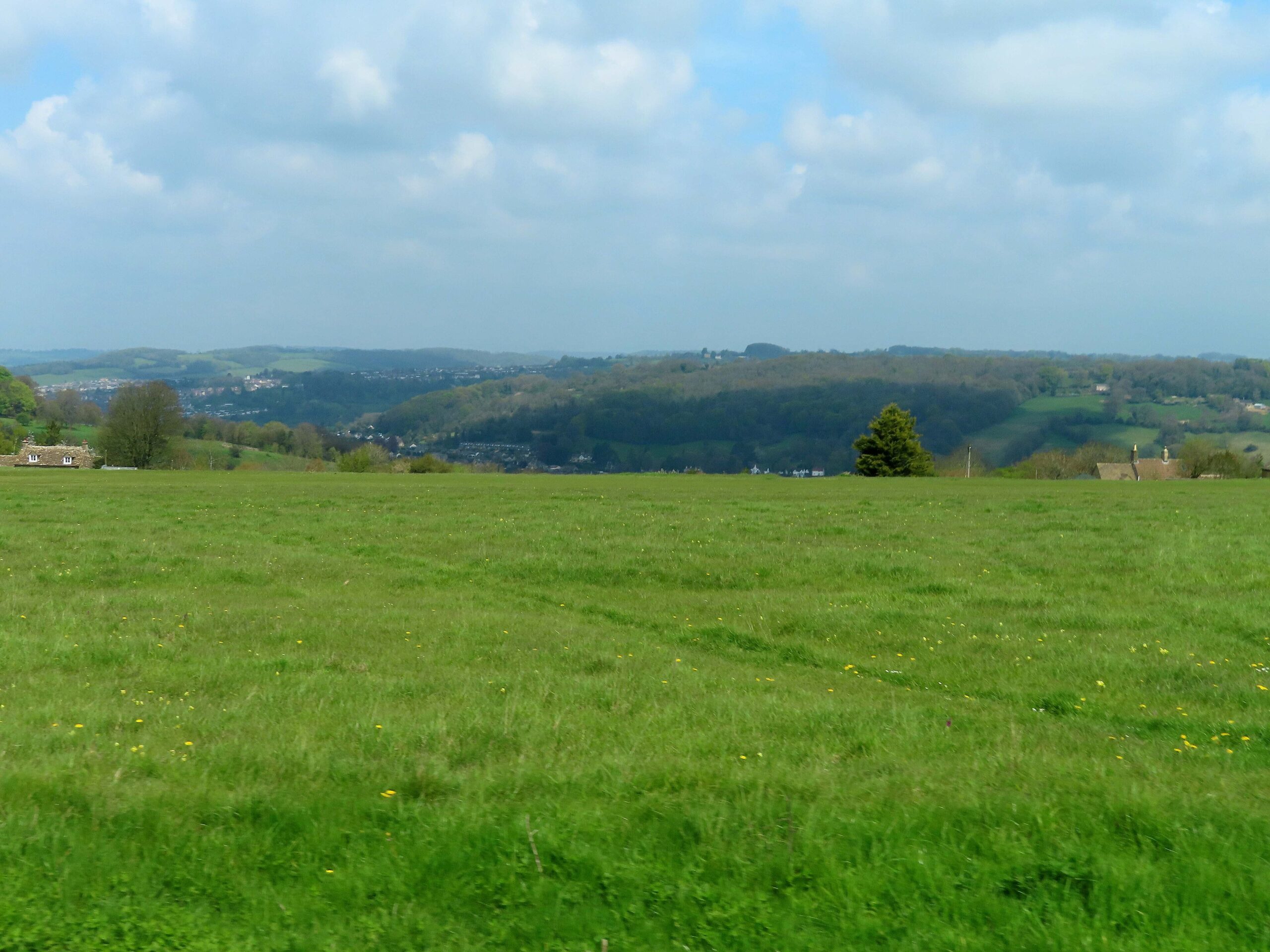 Open grassland on Minchinhampton Common with wooded valley sides and the rooftops of the Stroud valleys below under a cloudy spring sky
