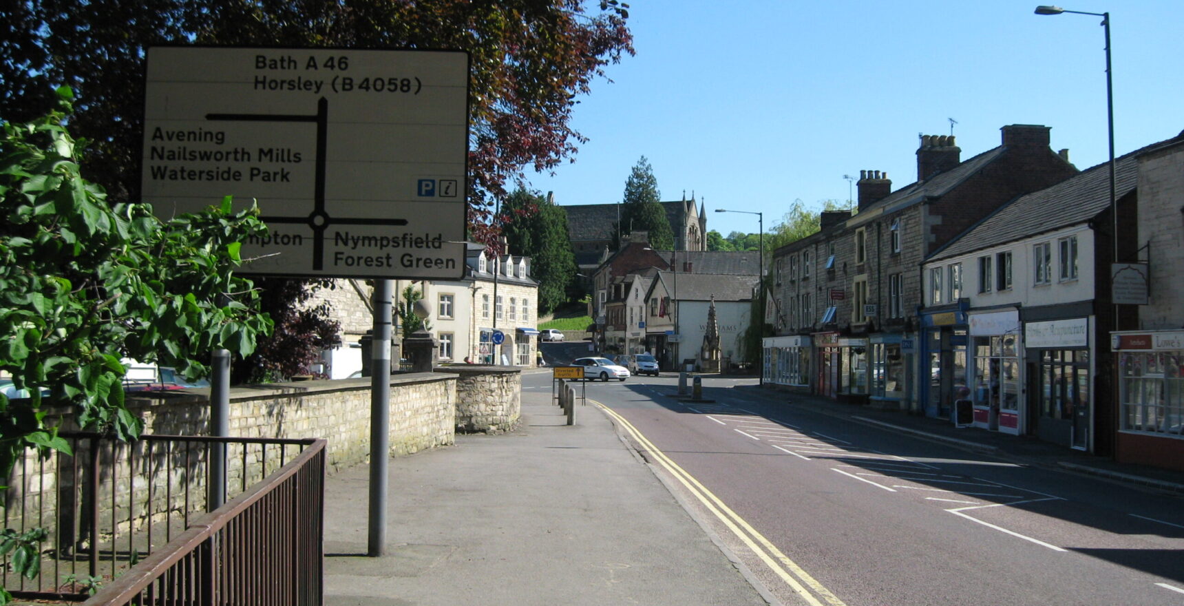 The road into Nailsworth town centre on a sunny morning, with a terrace of stone-built shops on the right and the town's clock tower at the junction ahead