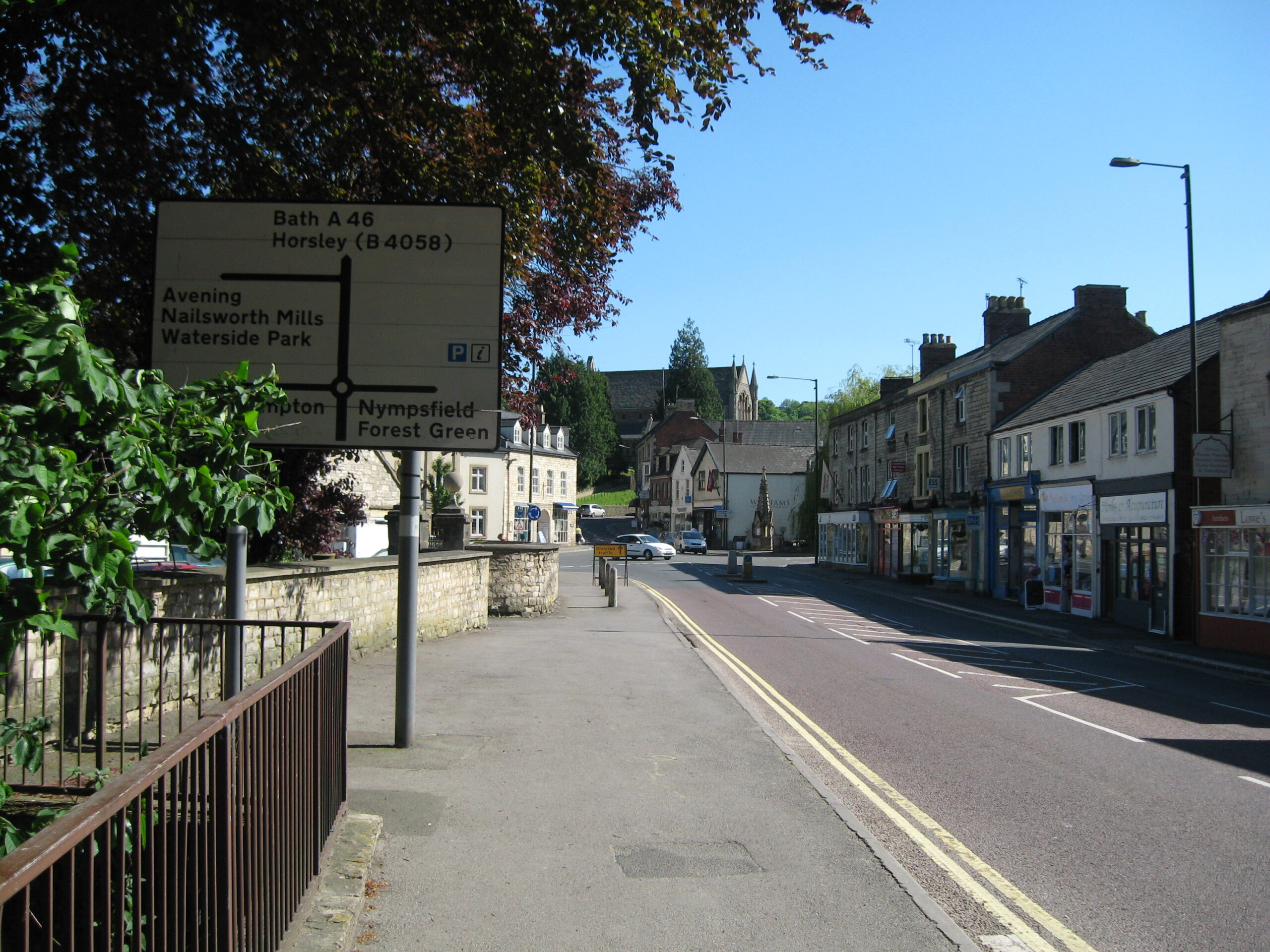 The road into Nailsworth town centre on a sunny morning, with a terrace of stone-built shops on the right and the town's clock tower at the junction ahead