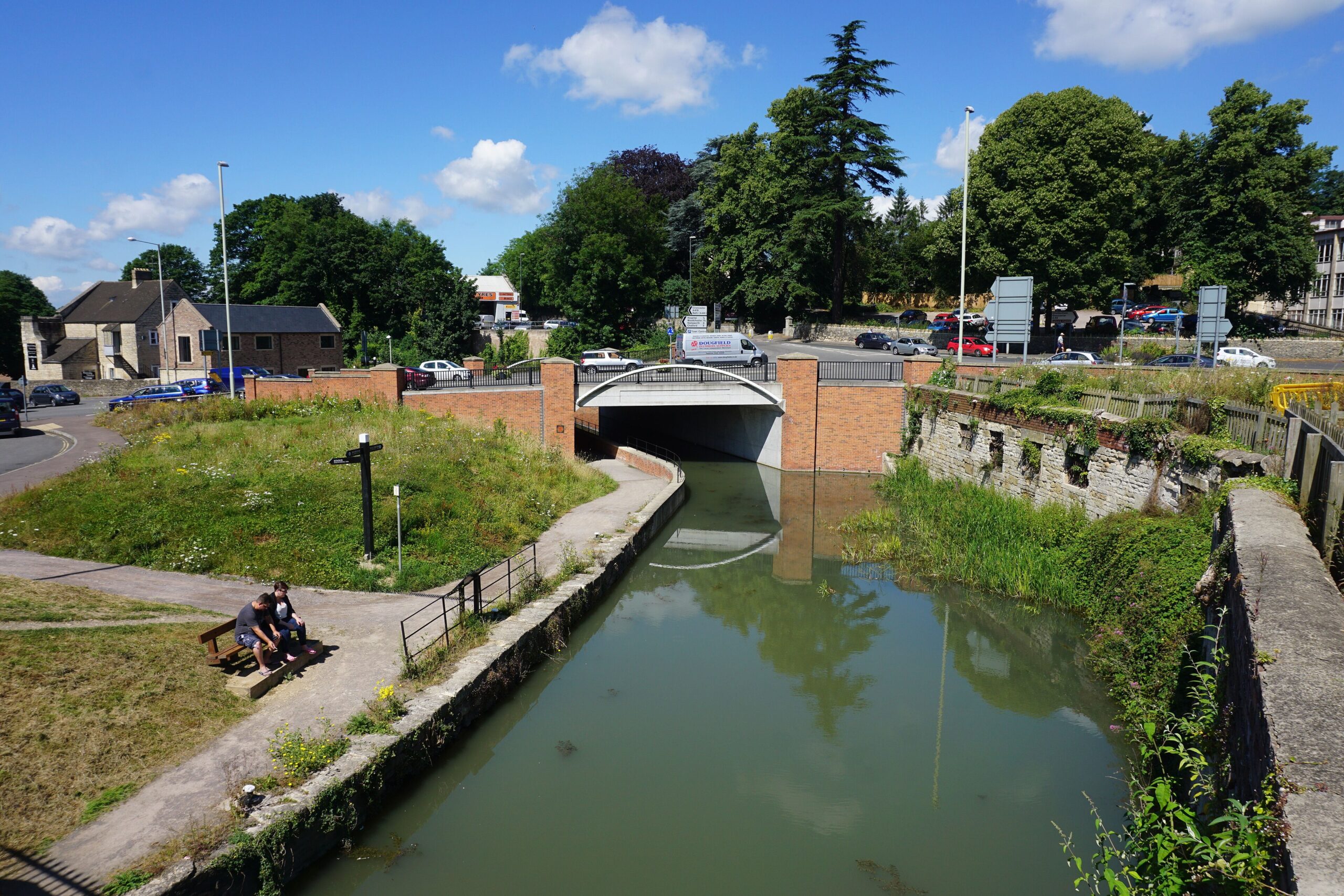 The restored Stroudwater Canal in Stroud running under a road bridge, with a towpath, a bench and a couple sitting beside the water