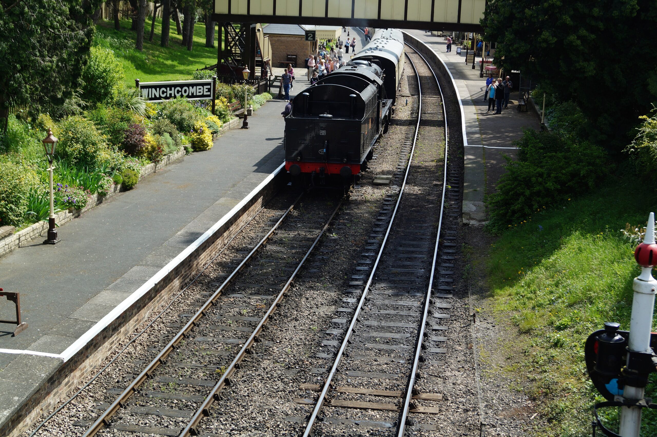 A black steam locomotive with maroon coaches standing at Winchcombe station on the Gloucestershire Warwickshire Railway, with the Winchcombe nameboard, gas lamps and flower beds on the platform