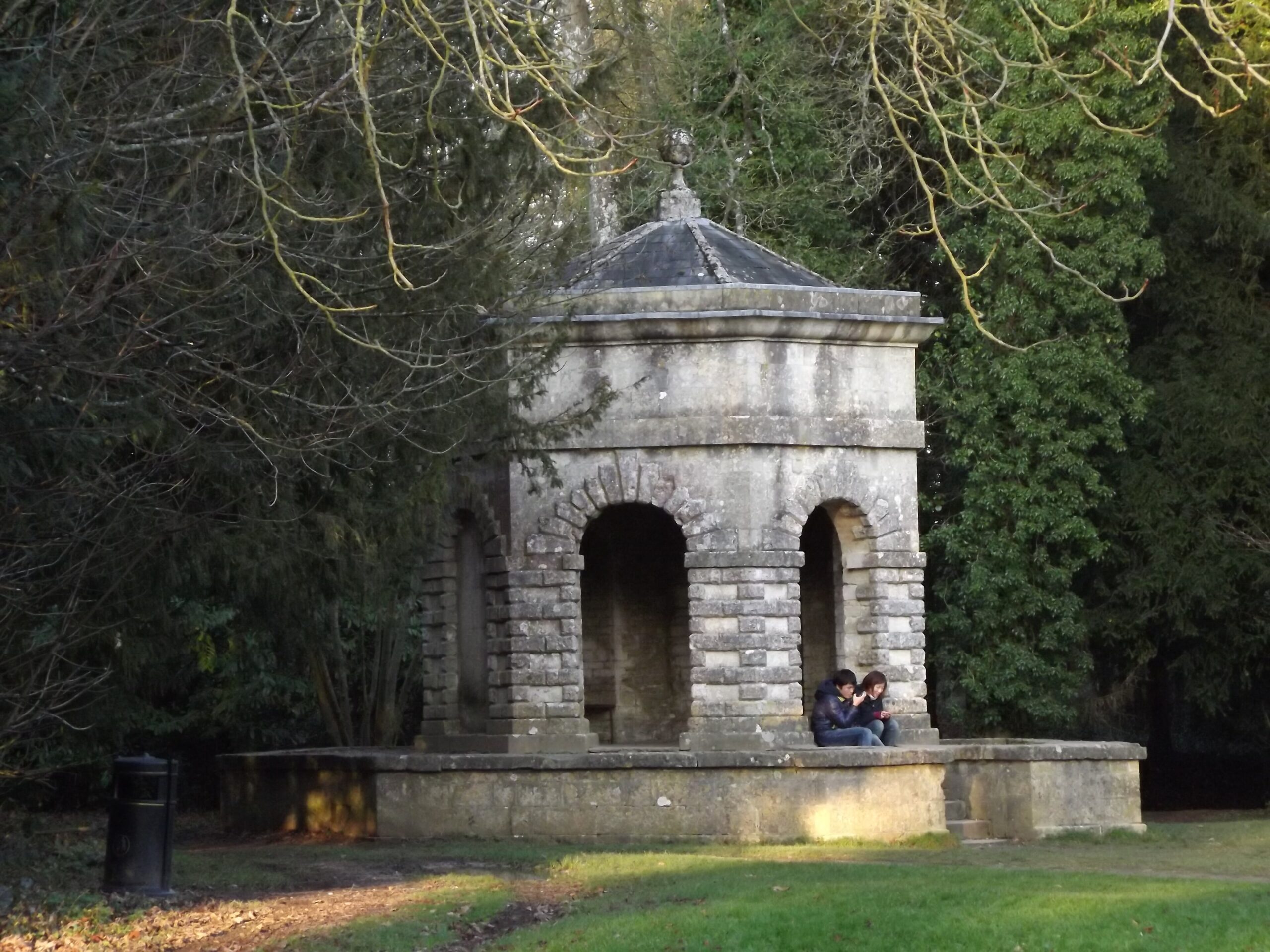 The Hexagon, a small eighteenth-century stone folly with arched openings, beside a path under trees in Cirencester Park