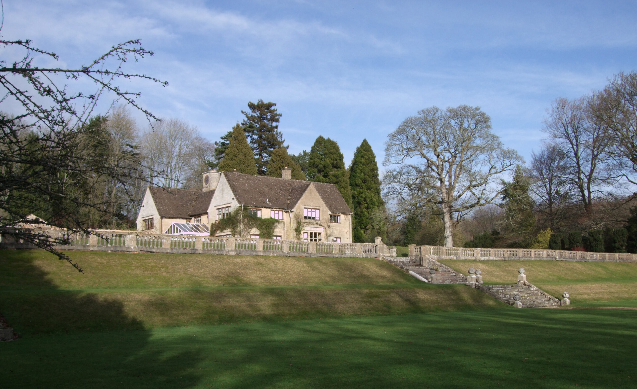 The stone house at Colesbourne Park above a terraced lawn with a balustrade, under a blue early-spring sky