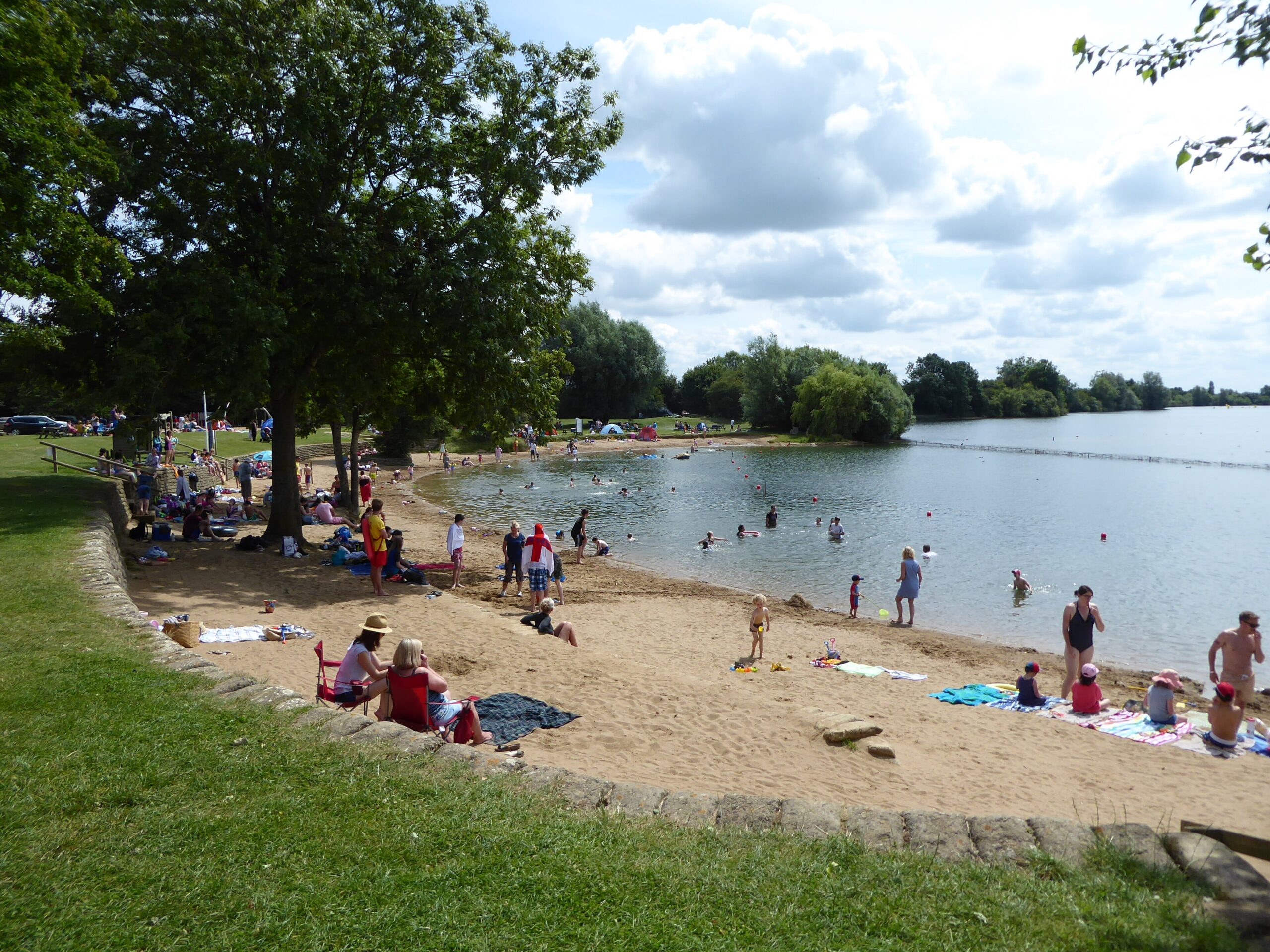Families on the sandy artificial beach at Cotswold Country Park and Beach, with swimmers and paddlers in the lake beyond