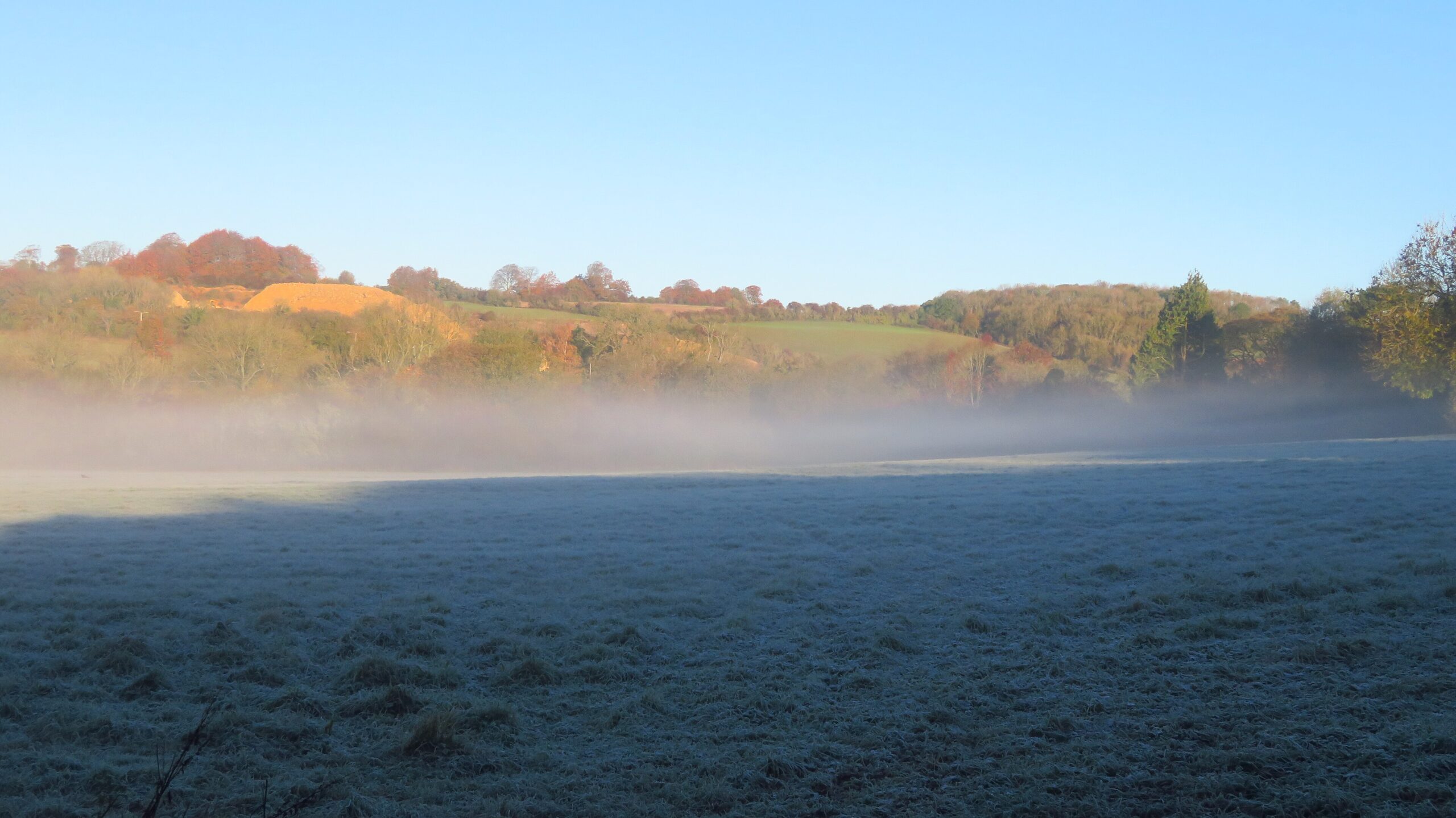Early morning mist lying over a frosted field in the valley at Syreford, with low sun on the wooded hillside beyond