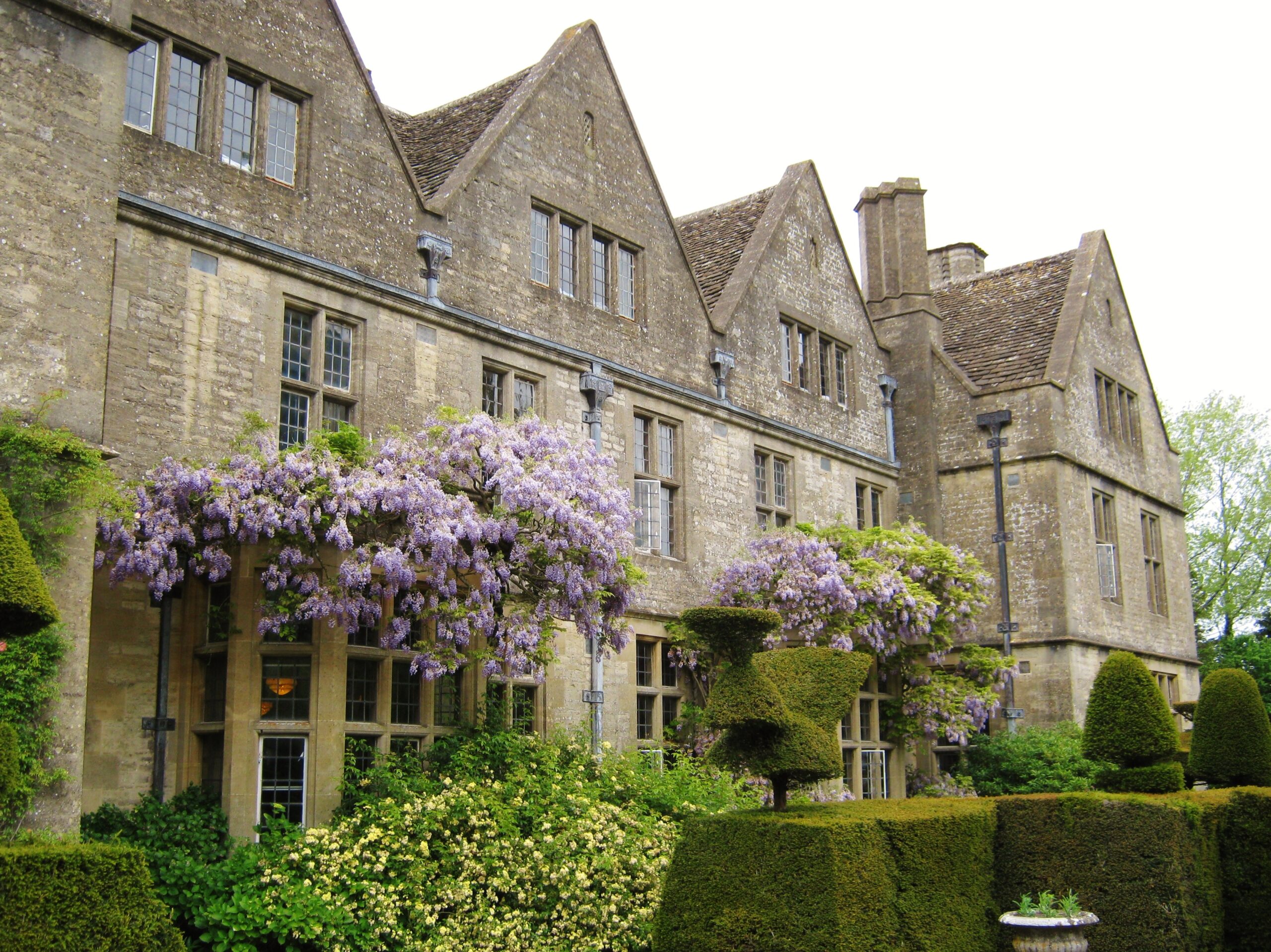 The gabled stone front of Rodmarton Manor with wisteria in flower and clipped topiary hedges