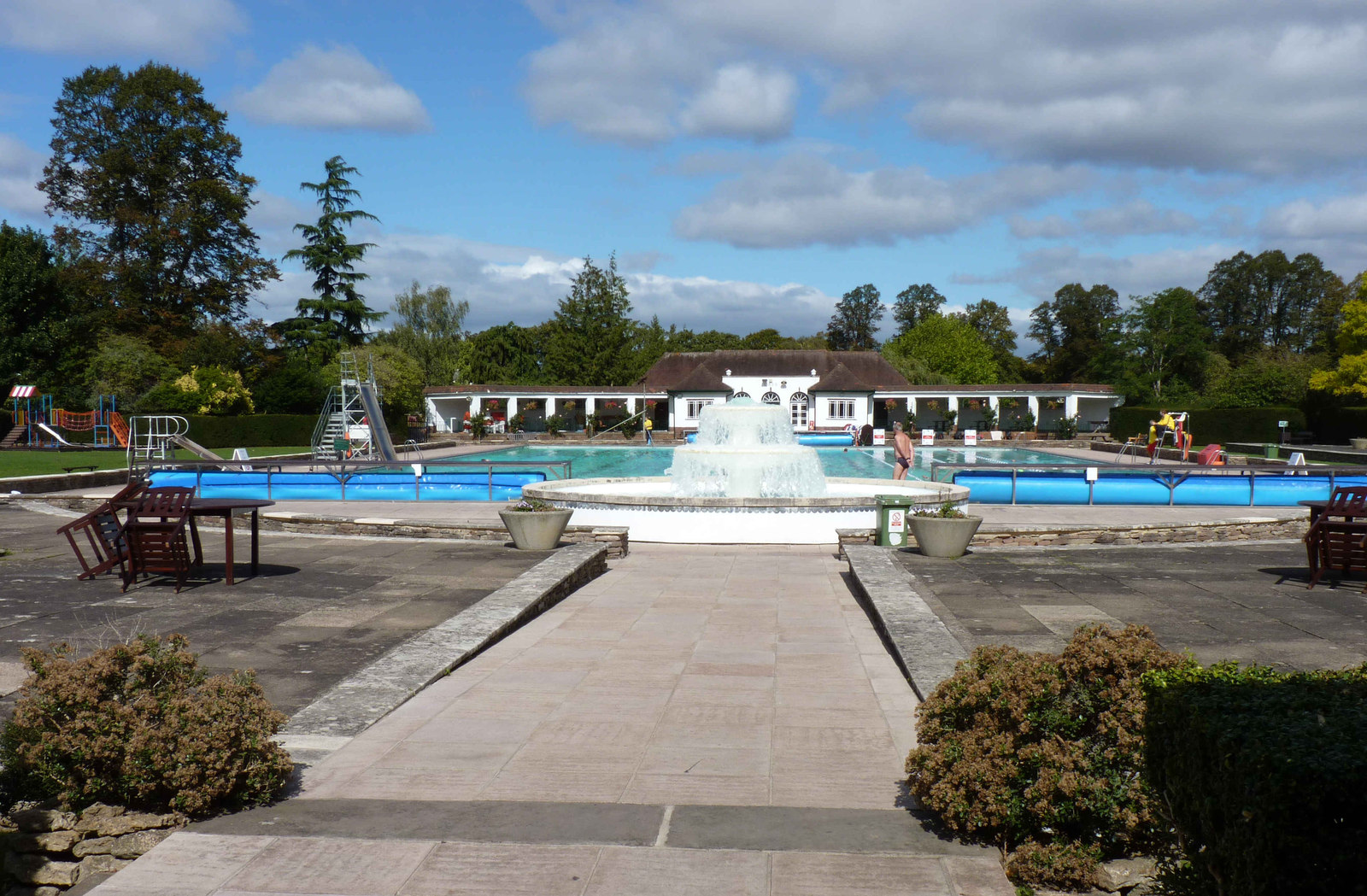 The fountain and paved terrace at Sandford Parks Lido, with the blue main pool, diving boards and a white pavilion behind